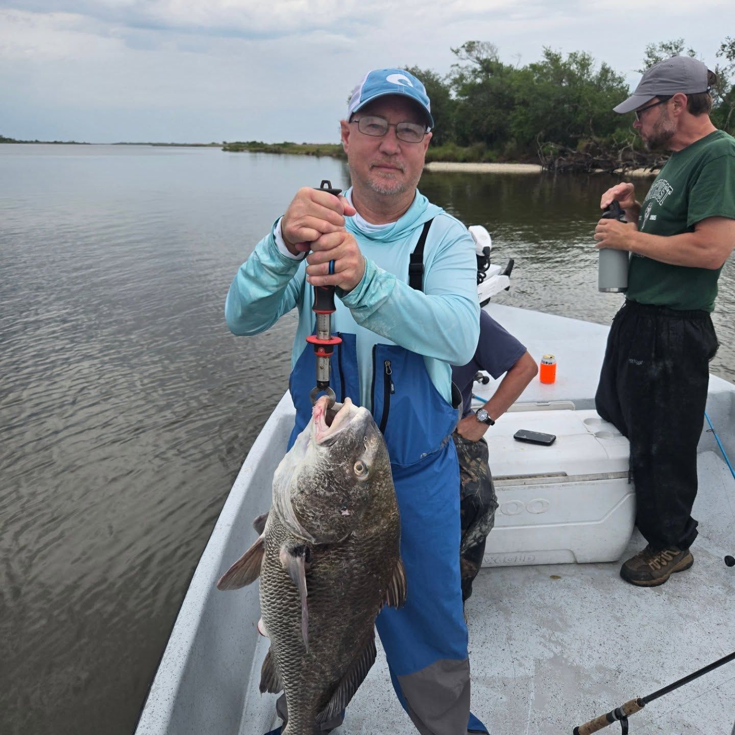Man holding a large fish on a boat with two others beside him on calm water.