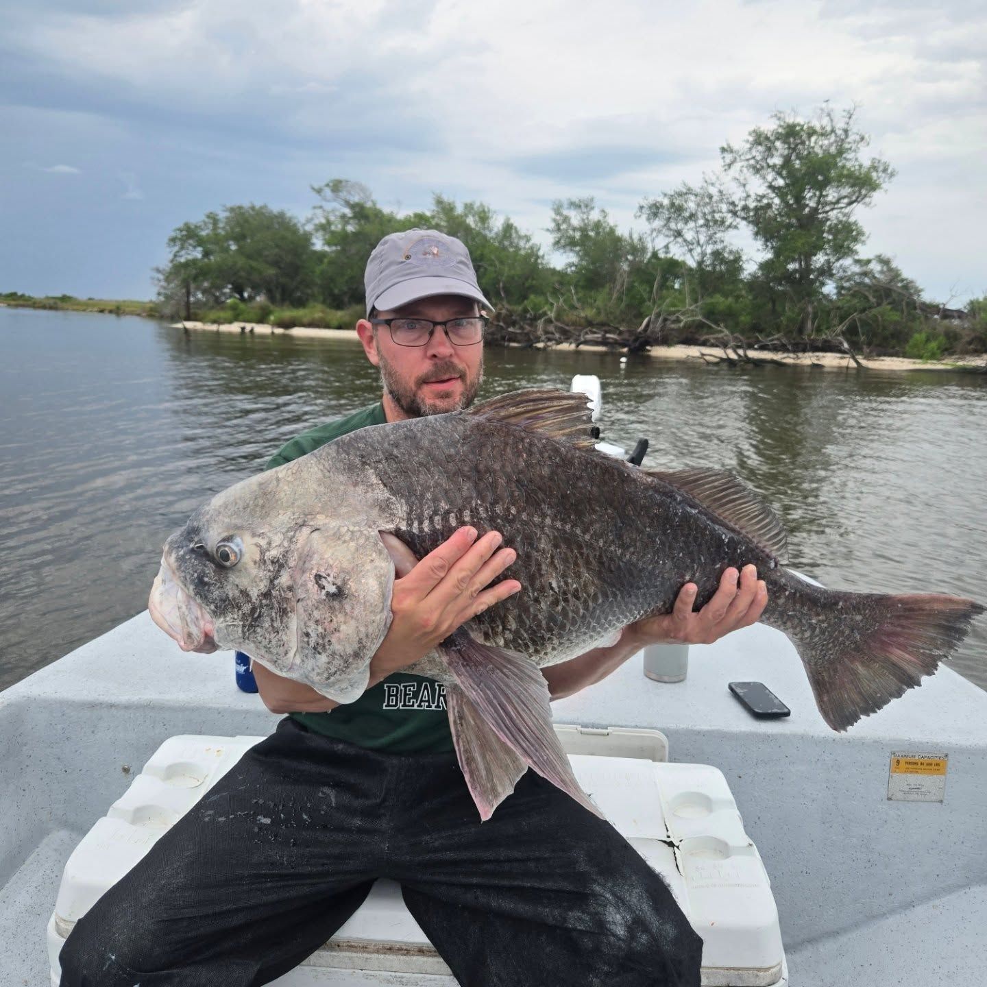 Man on a boat holding a large fish in a calm river or bay, with trees in the background.