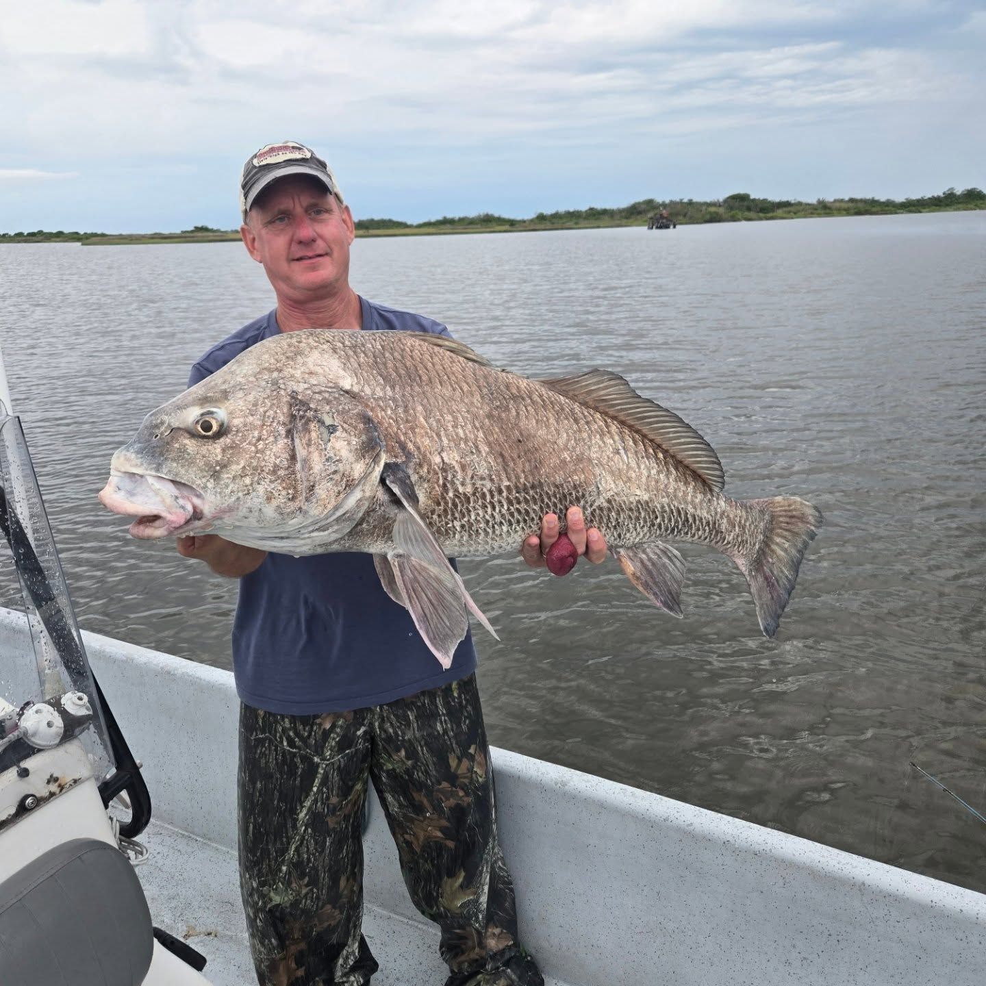 Man holding a large fish on a boat in calm water under an overcast sky