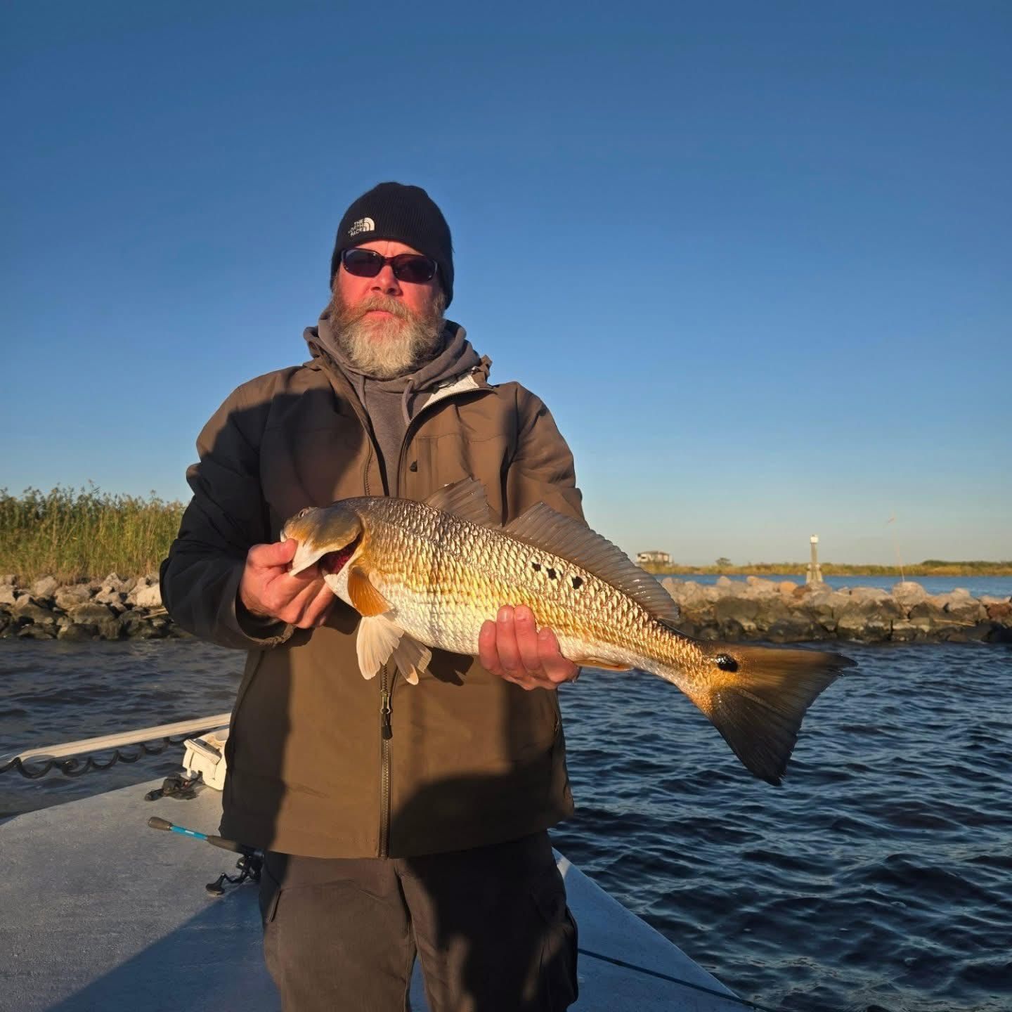 Man holding a large fish on a dock at sunset, wearing winter gear and sunglasses
