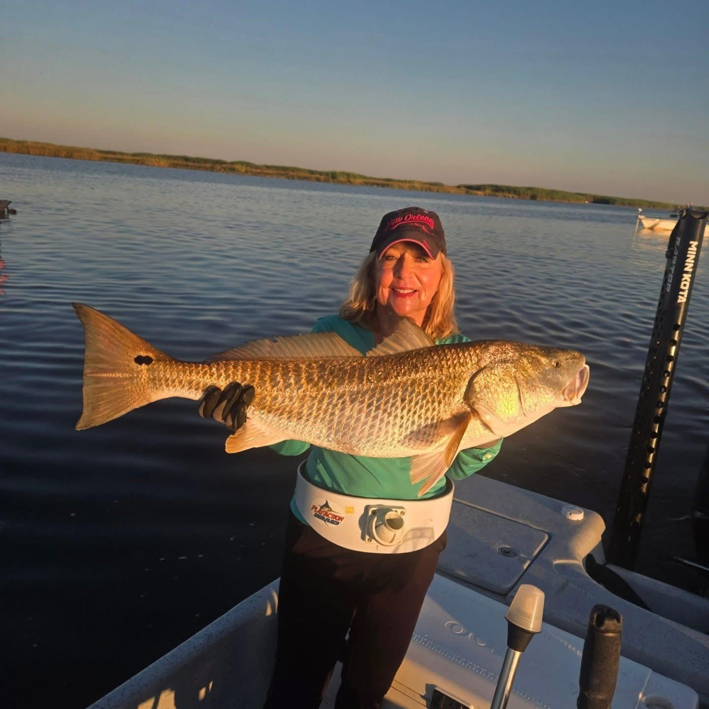 Woman holding a large fish on a boat at sunset, with calm water and shoreline in the background