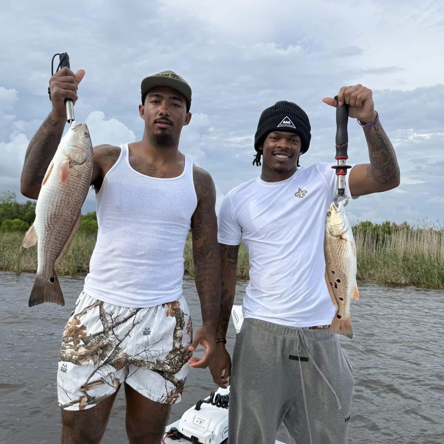 Two men on a boat hold up large fish in a marsh under a cloudy sky