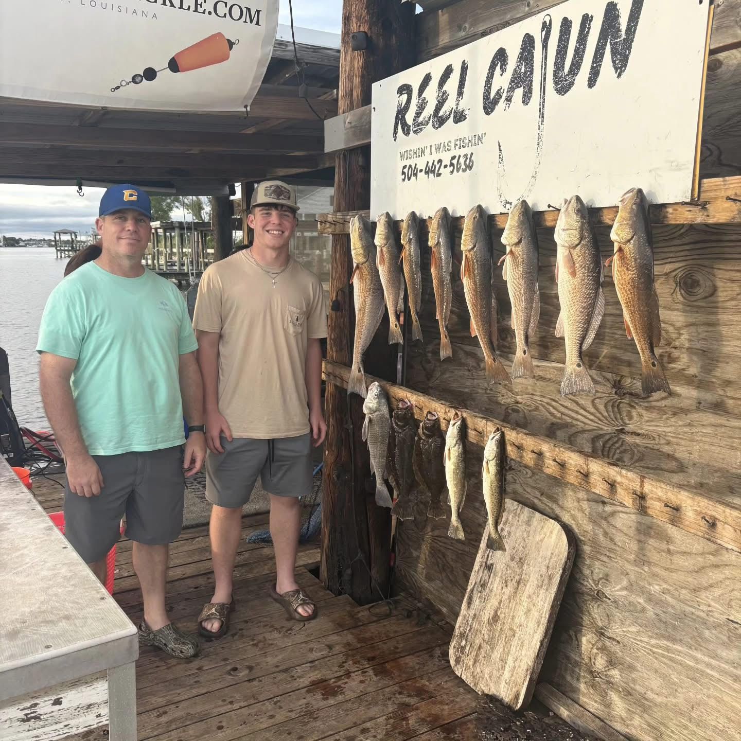 Two men standing beside a wall display of hanging fish under a sign reading “Reel Cajun”