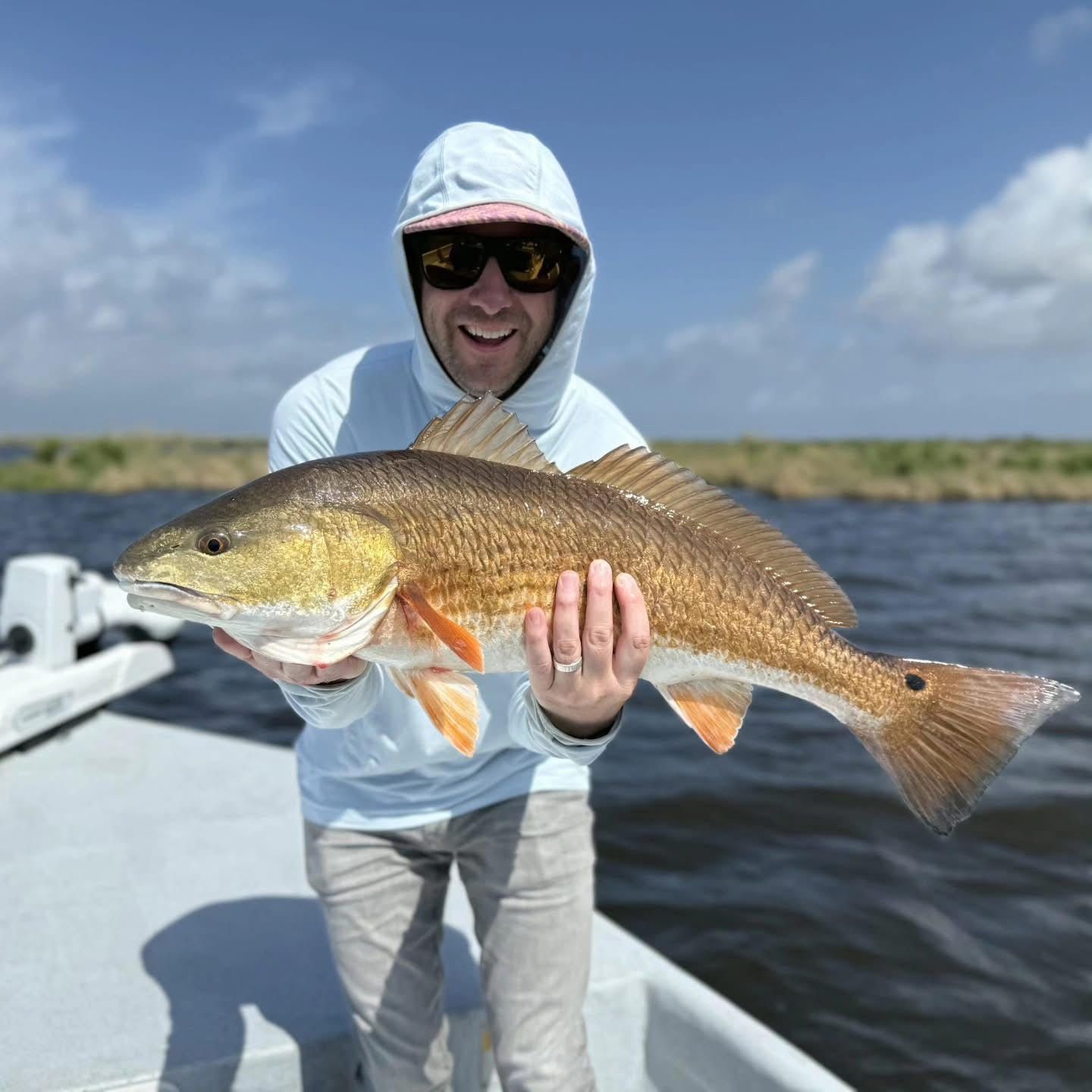 Angler in a hooded jacket holding a large fish on a boat under a sunny blue sky