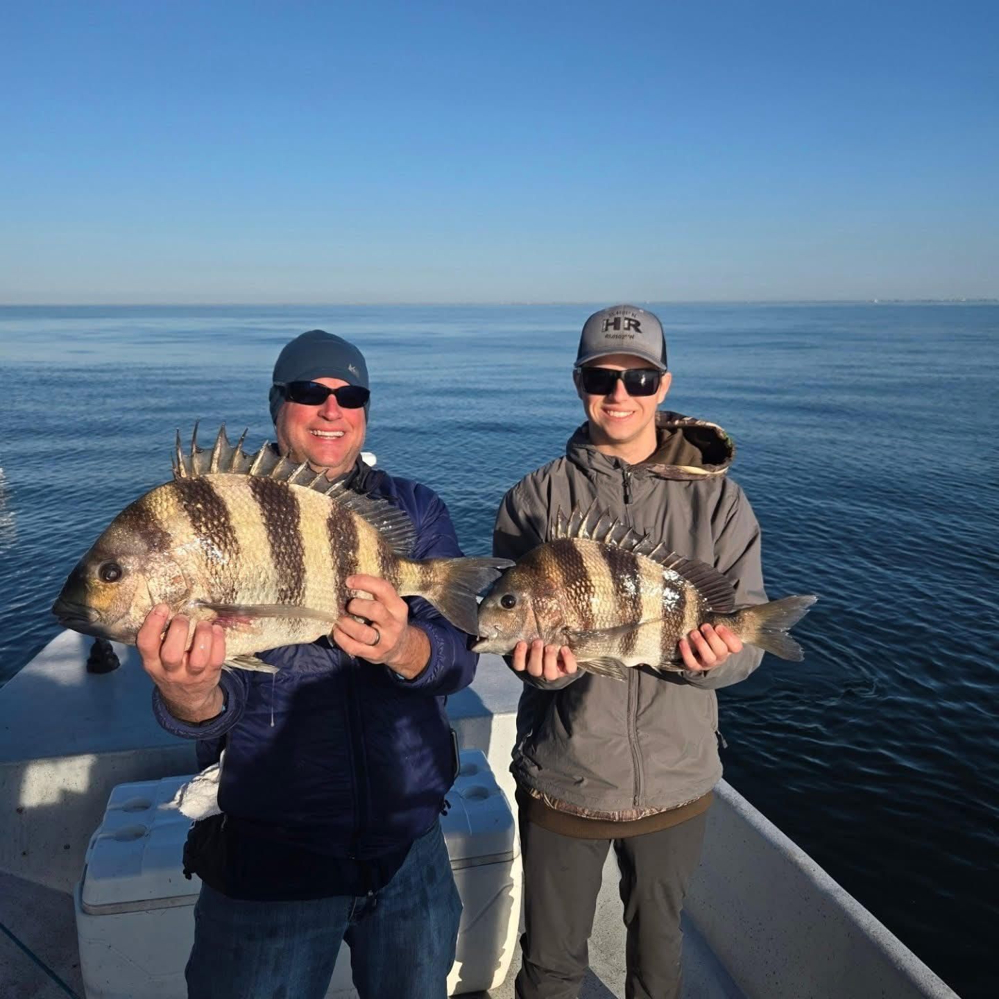Two people on a boat holding large striped fish with the ocean behind them