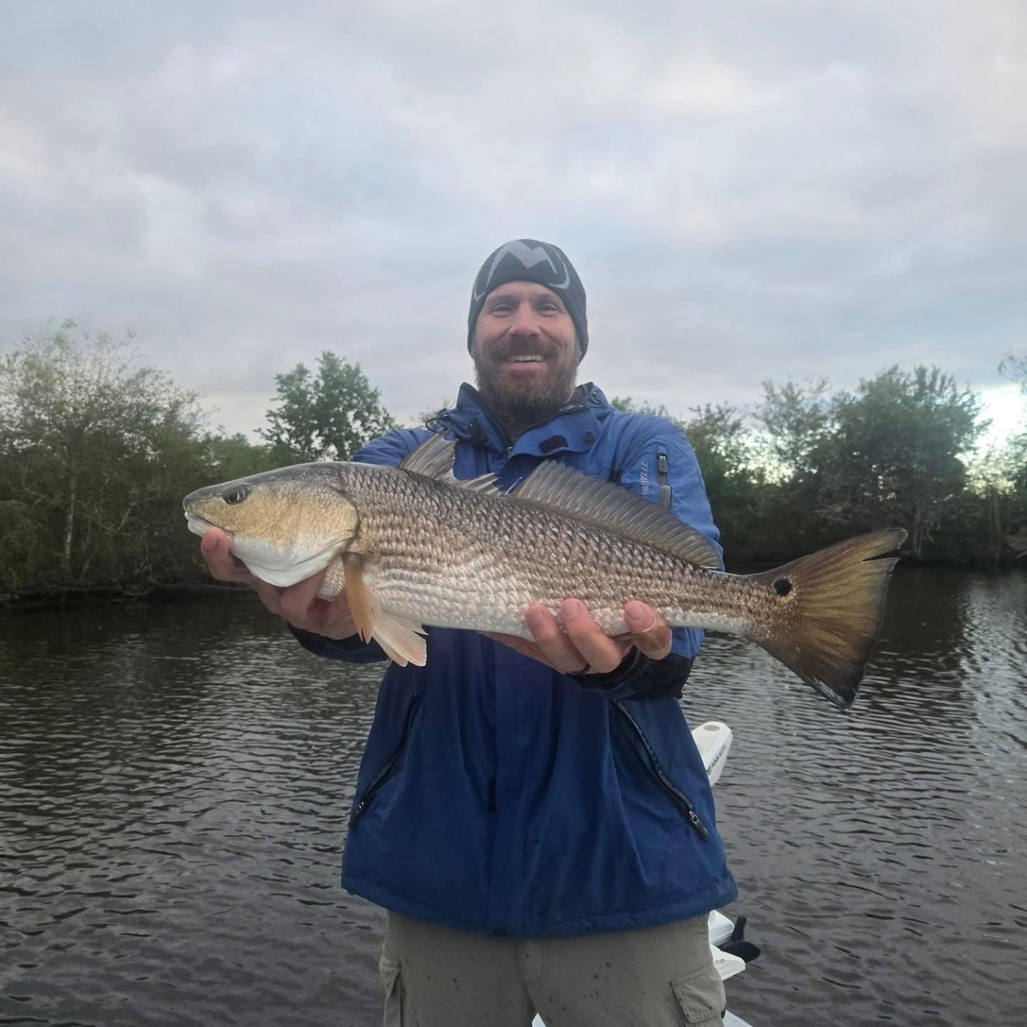 Angler in blue jacket holding a large fish by a calm river under cloudy sky