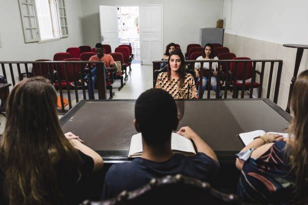 A courtroom scene with people seated at tables, looking toward a witness.