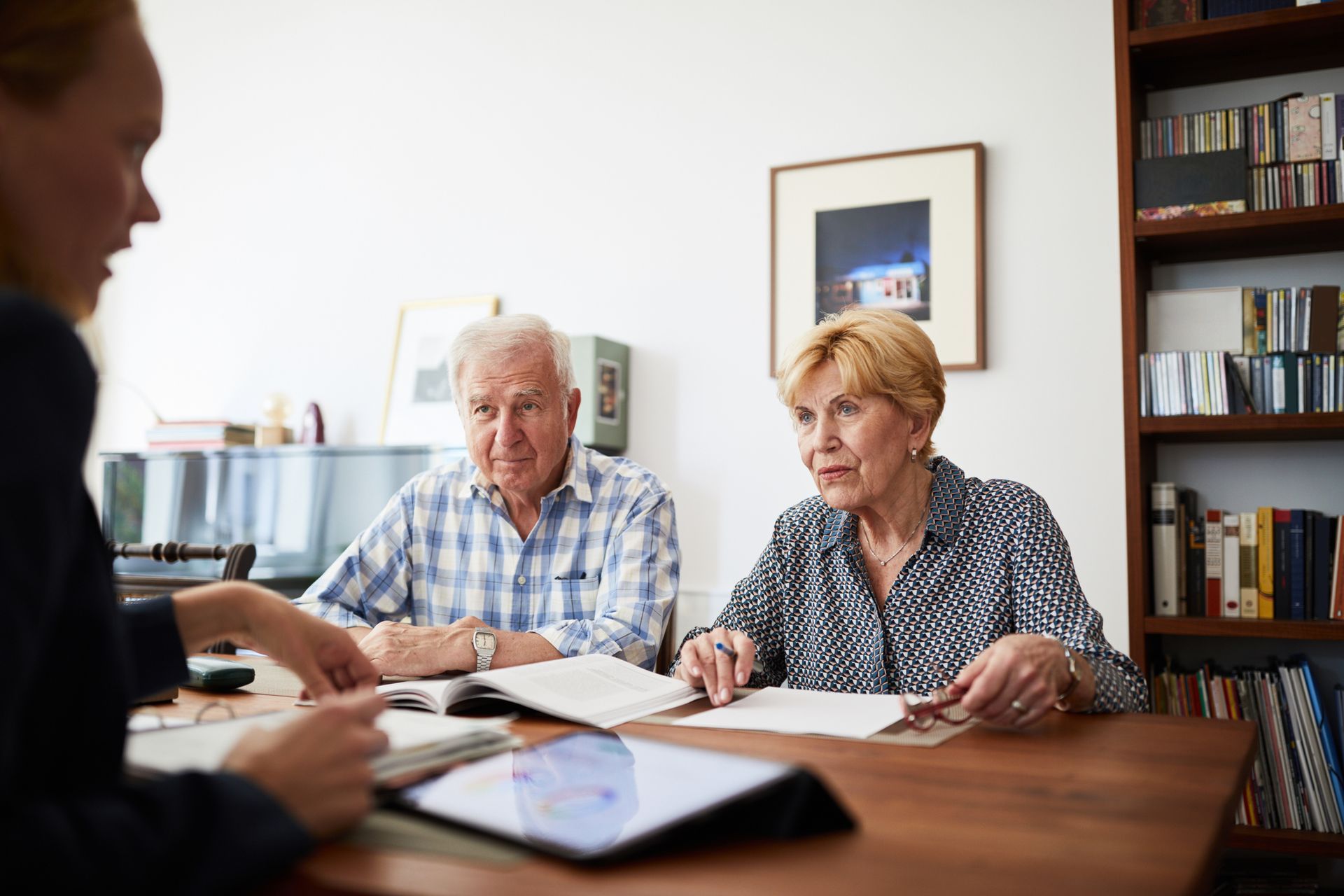 Woman with older couple at a table, looking at documents and a tablet. Interior setting with bookshelf.