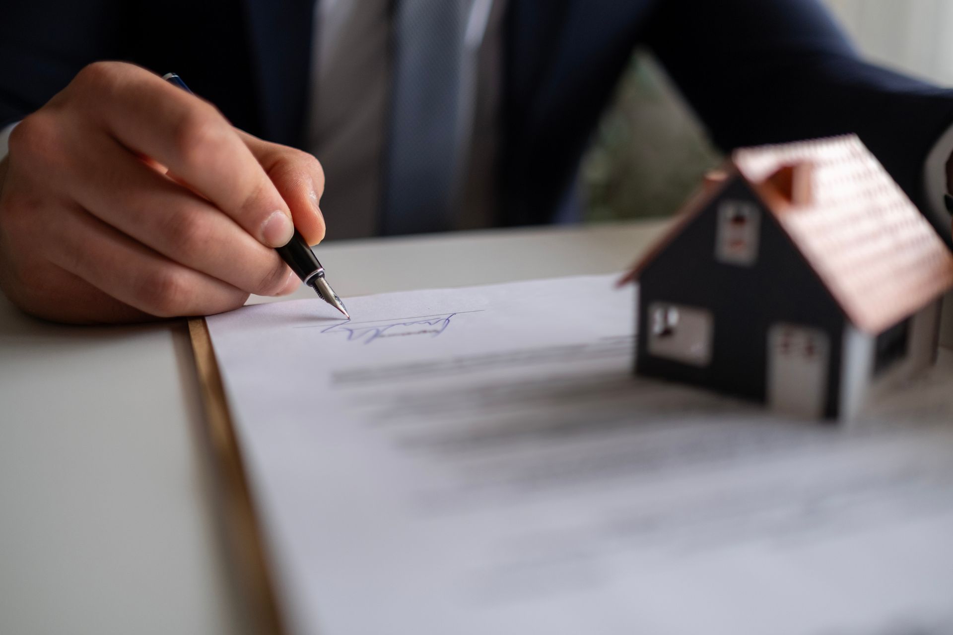 Person signing a document with a pen next to a miniature house model.