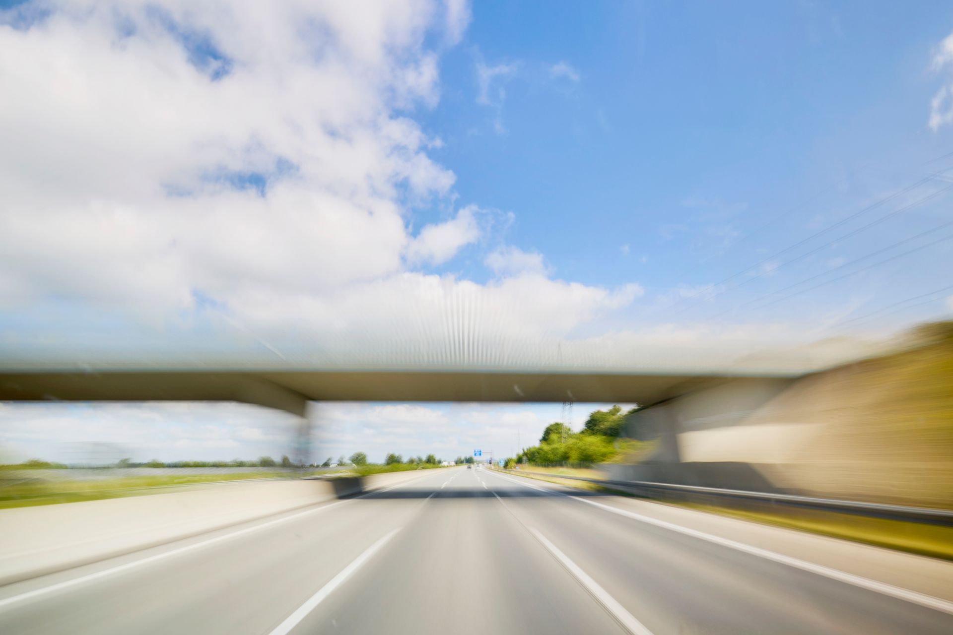 Blurred view of highway passing under a bridge, with blue sky and clouds overhead.