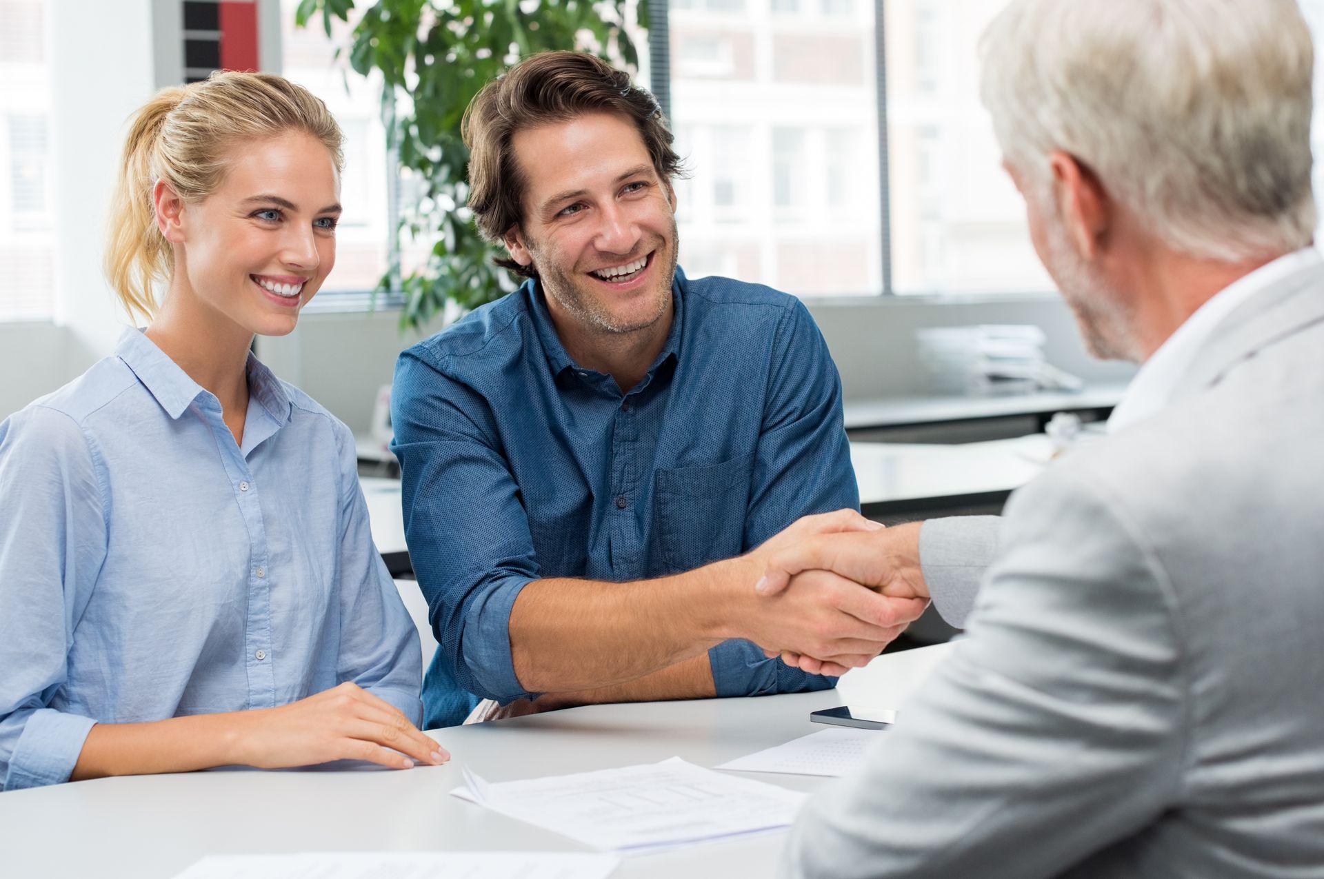 Couple smiles, shaking hands with a person in a light gray suit at a table. Bright office setting.