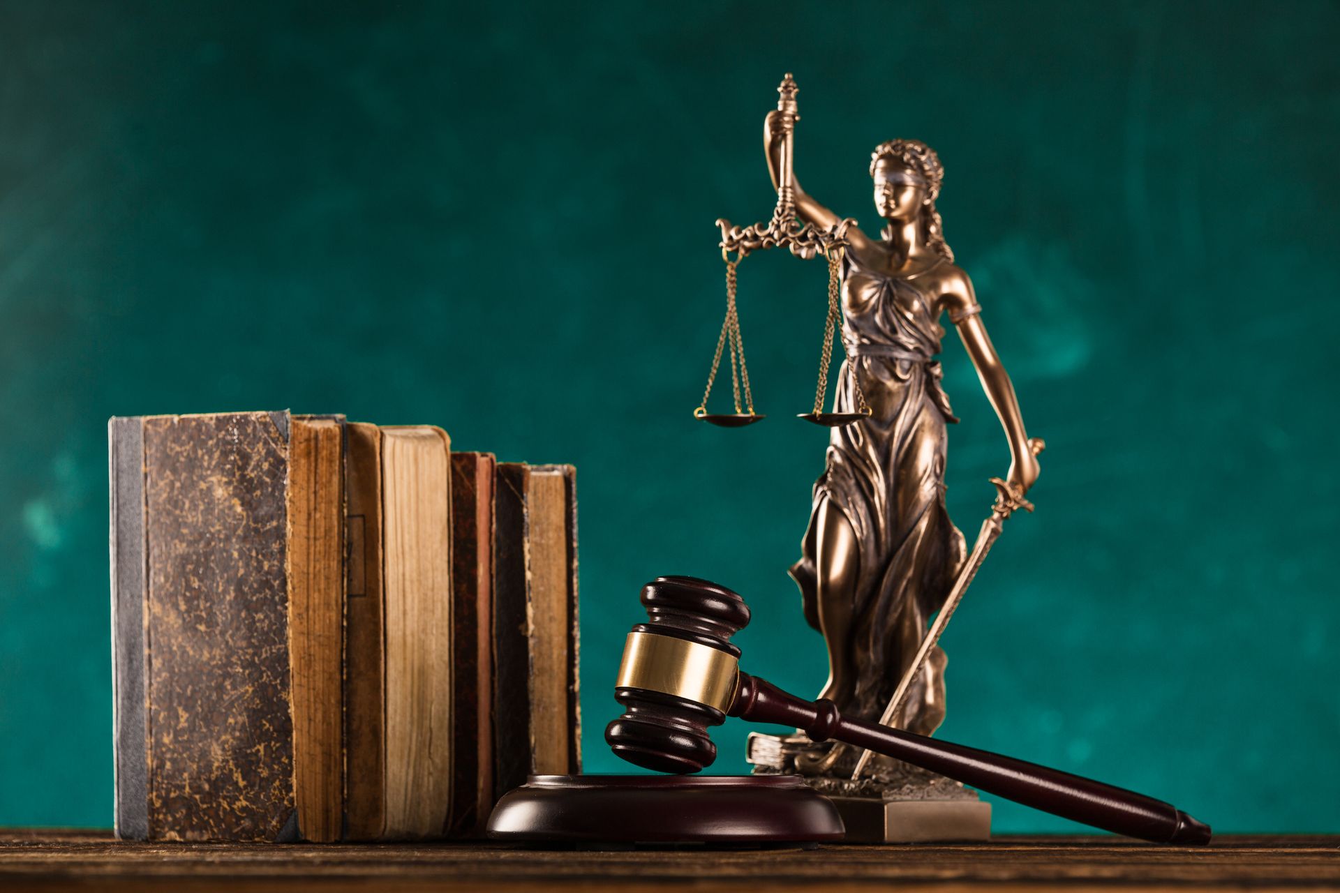 Statue of Lady Justice with scales and sword next to gavel and law books on wooden surface, green background.
