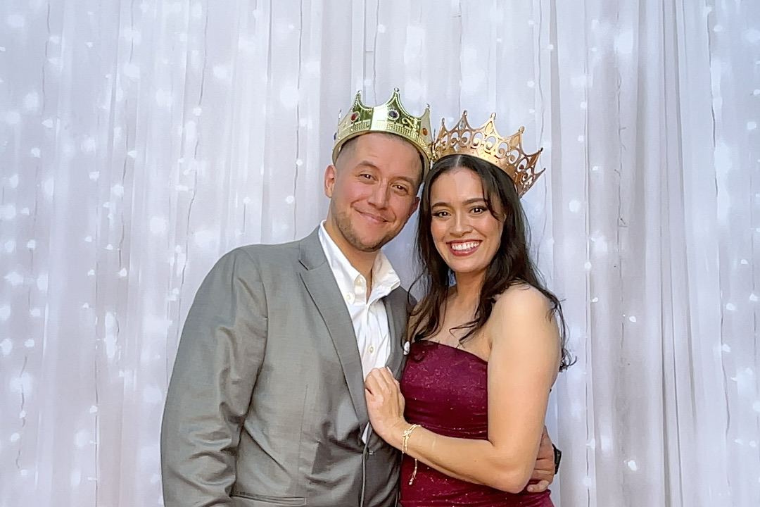 Couple wearing crowns, smiling, posing in front of a white backdrop with lights. The woman wears a burgundy dress.