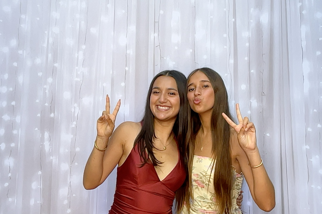 Couple wearing crowns, smiling, posing in front of a white backdrop with lights. The woman wears a burgundy dress.