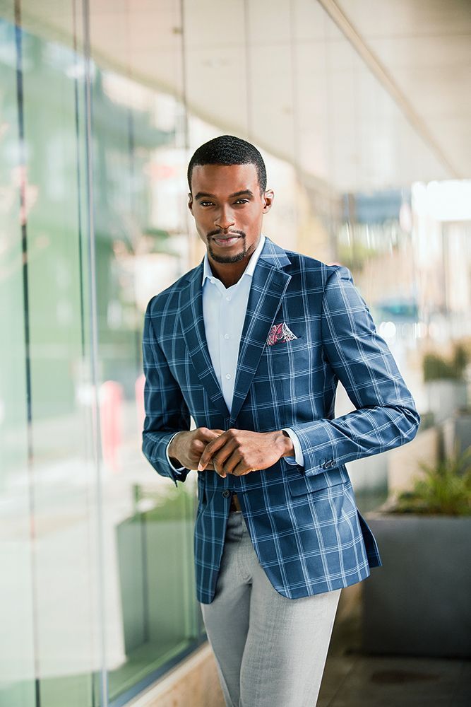 A man wearing a blue plaid jacket and grey pants is standing in front of a building.