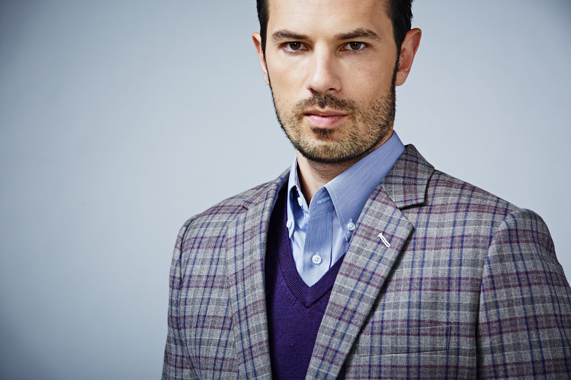 A man with a beard is wearing a plaid jacket and a purple vest.
