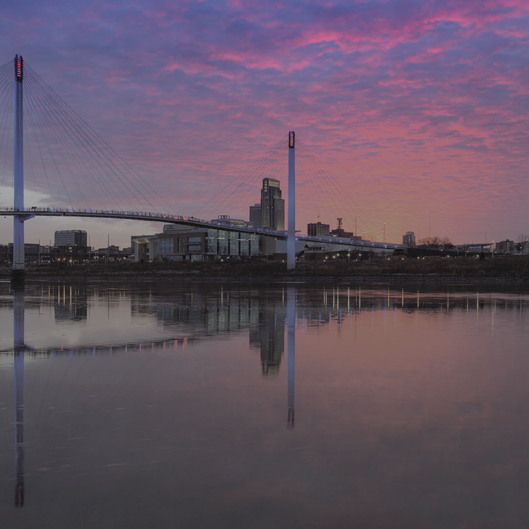 Bridge and skyline reflected in calm water at sunset; pink and purple sky.