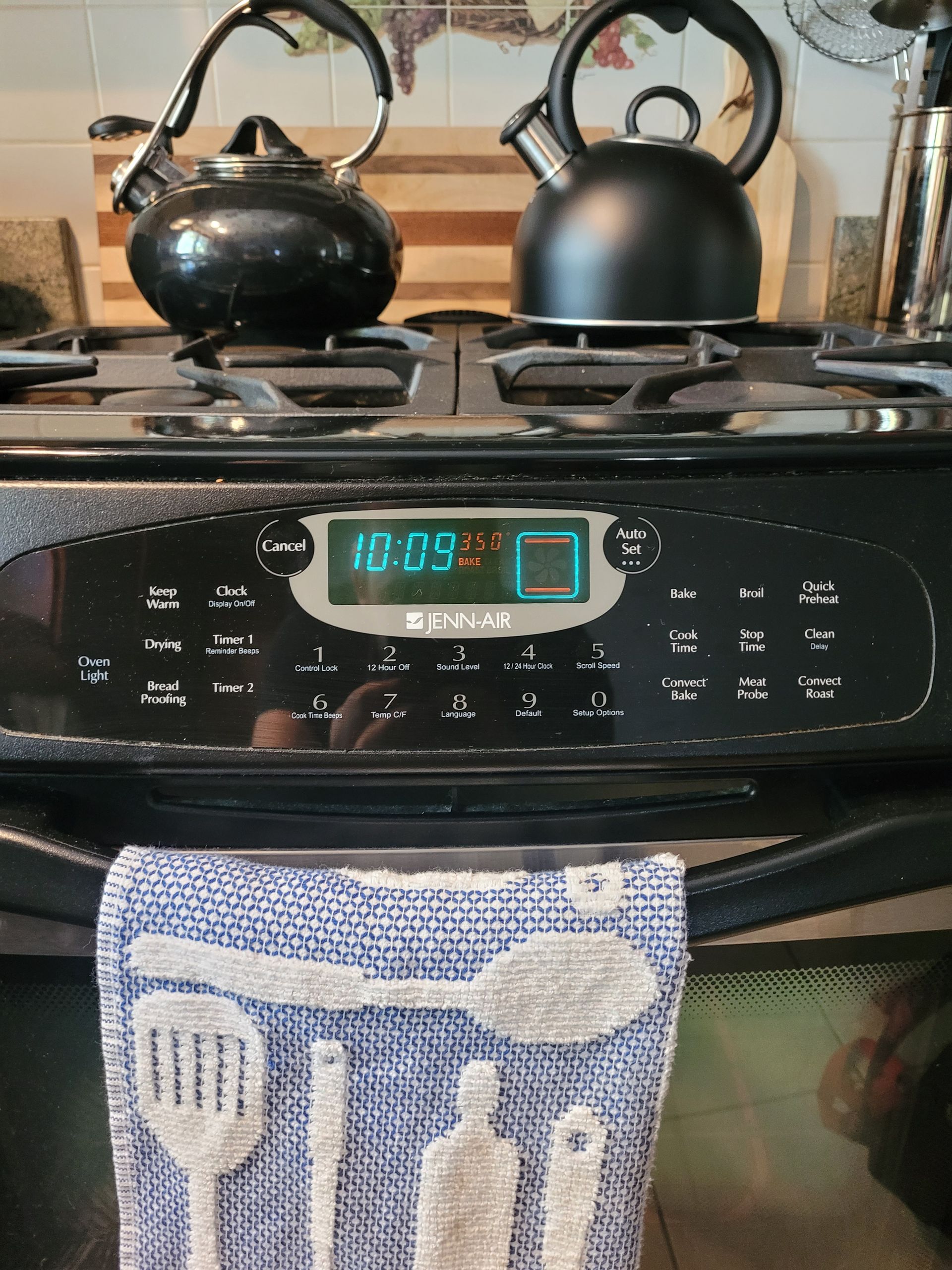 Two black tea kettles sit on a black stove, with a digital display reading 10:09 and a kitchen towel hanging in front.