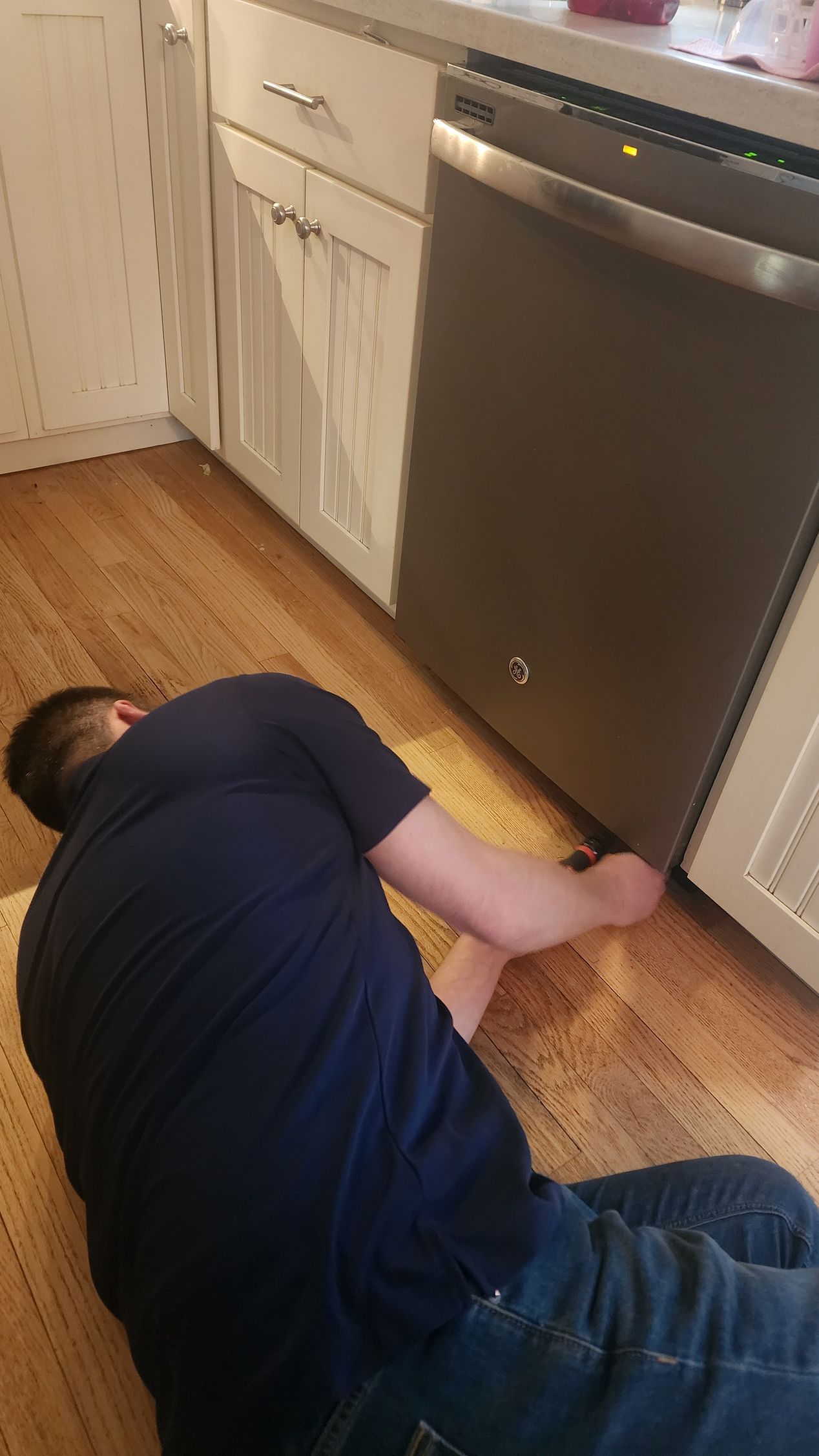 A person in a dark blue shirt kneeling on a kitchen floor, reaching underneath a stainless steel dishwasher.