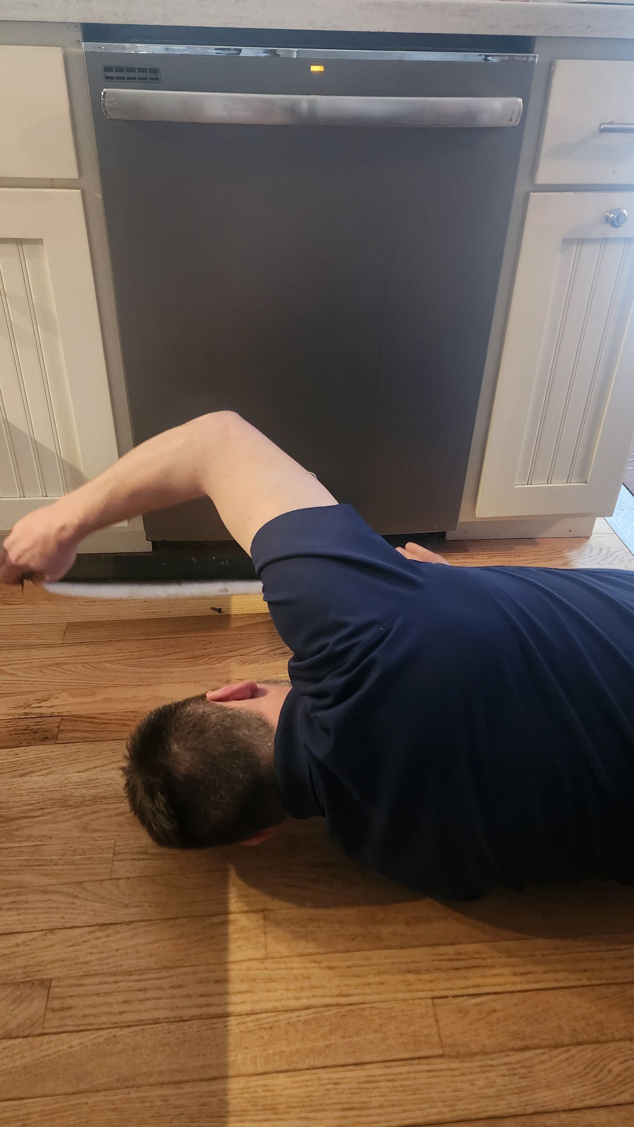 A person wearing a dark blue shirt lying on a wood floor, reaching under a stainless steel dishwasher in a kitchen.