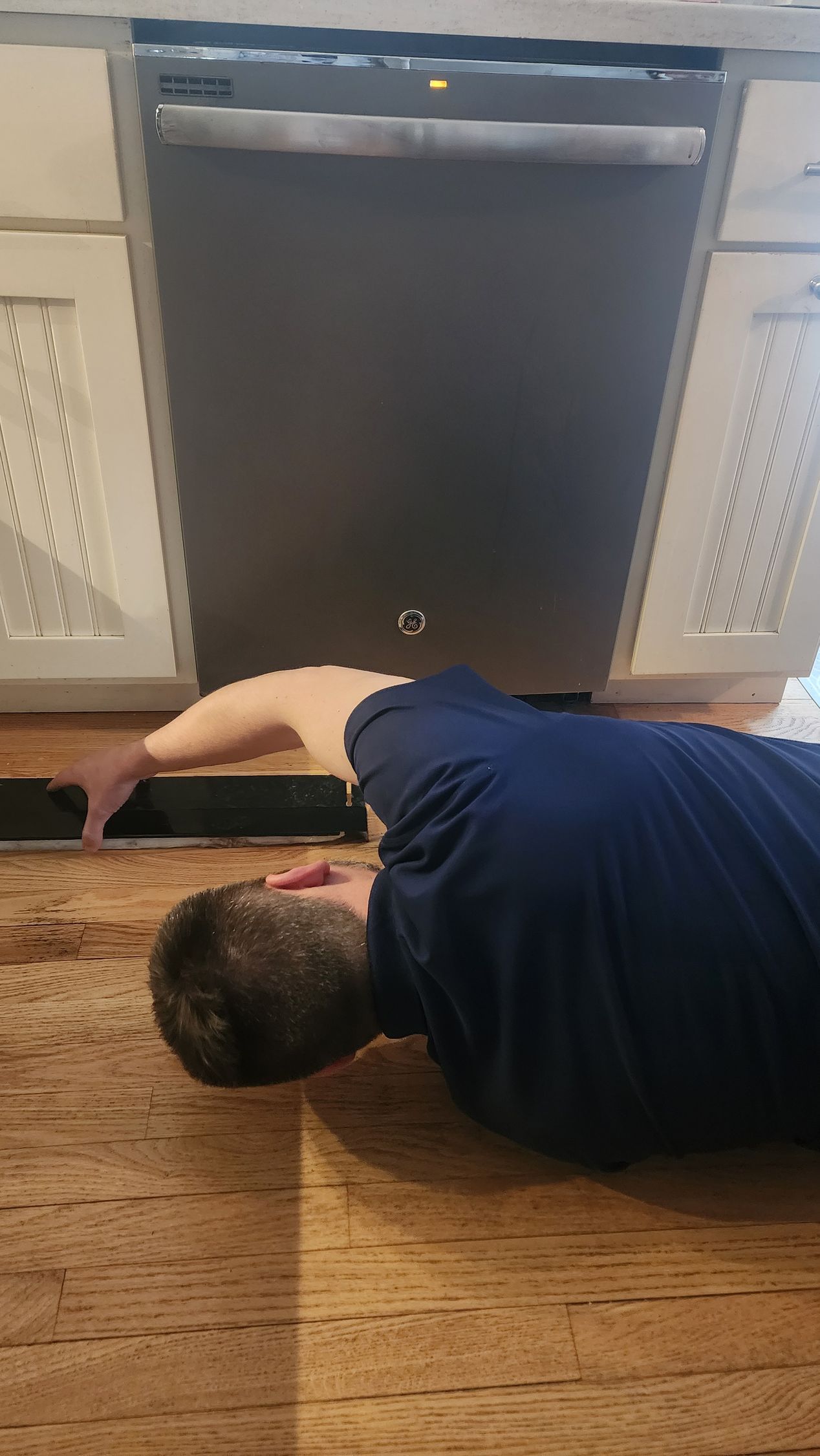 A person lies on a wooden floor, reaching under a stainless steel dishwasher to adjust the kickplate.