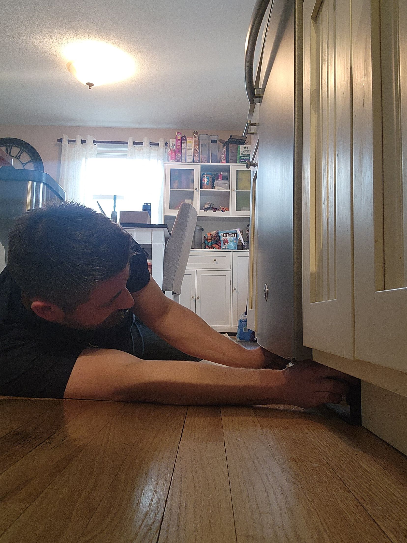 A person lies on a wooden floor, reaching under the base of a kitchen cabinet to perform a repair.