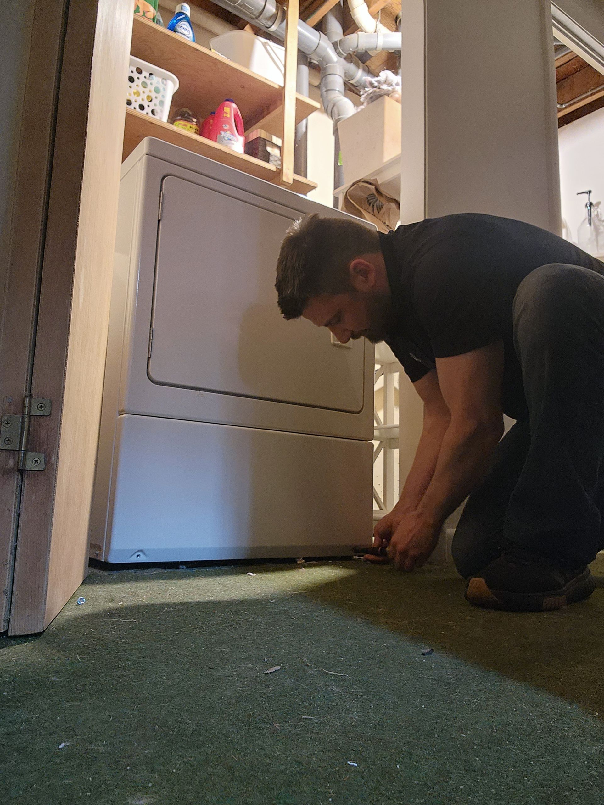 A person kneels on a green carpeted floor, working on the bottom of a white clothes dryer in a laundry room.