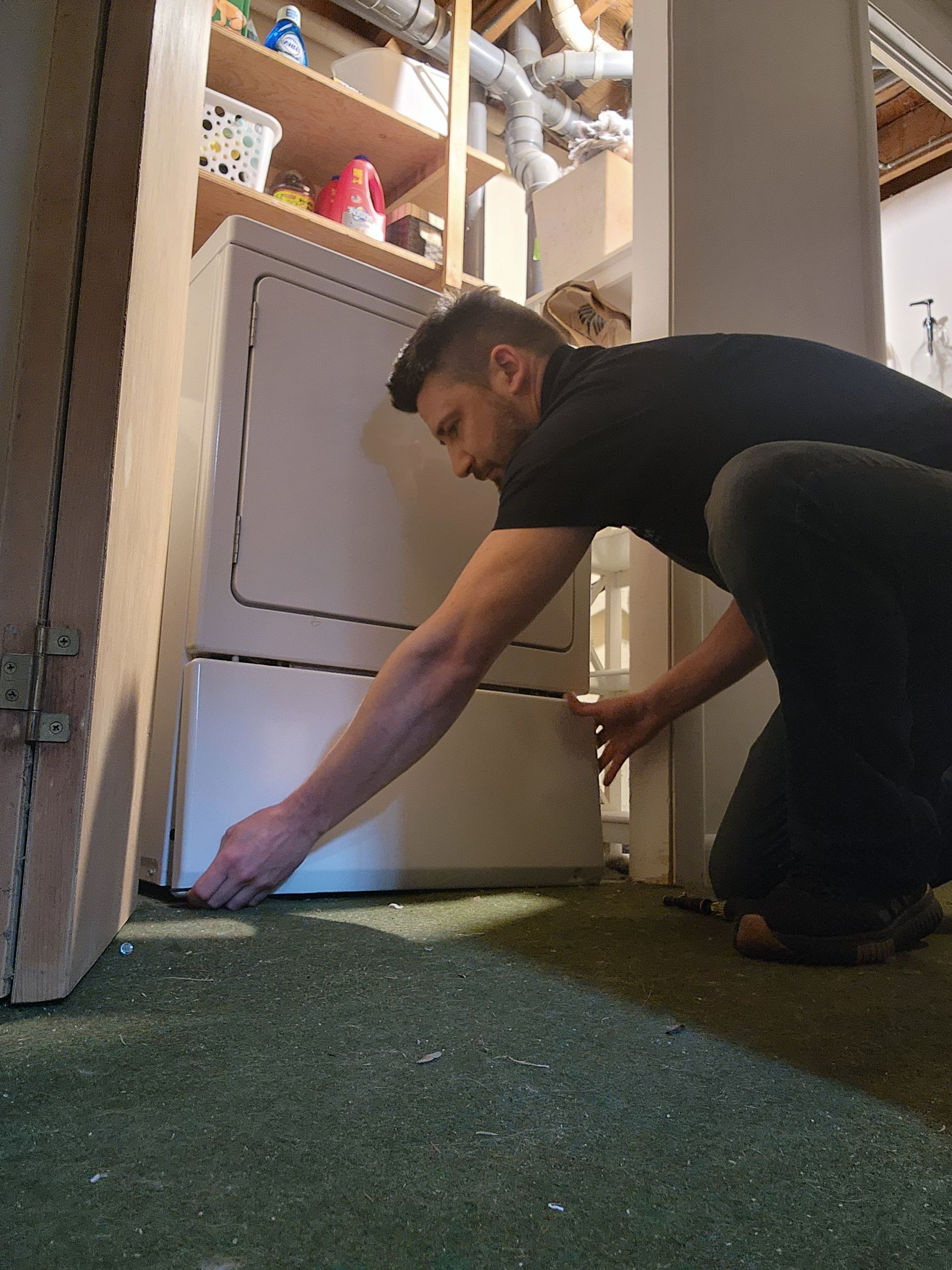 A person kneeling on a green carpeted floor while adjusting or installing a white appliance in a storage room.