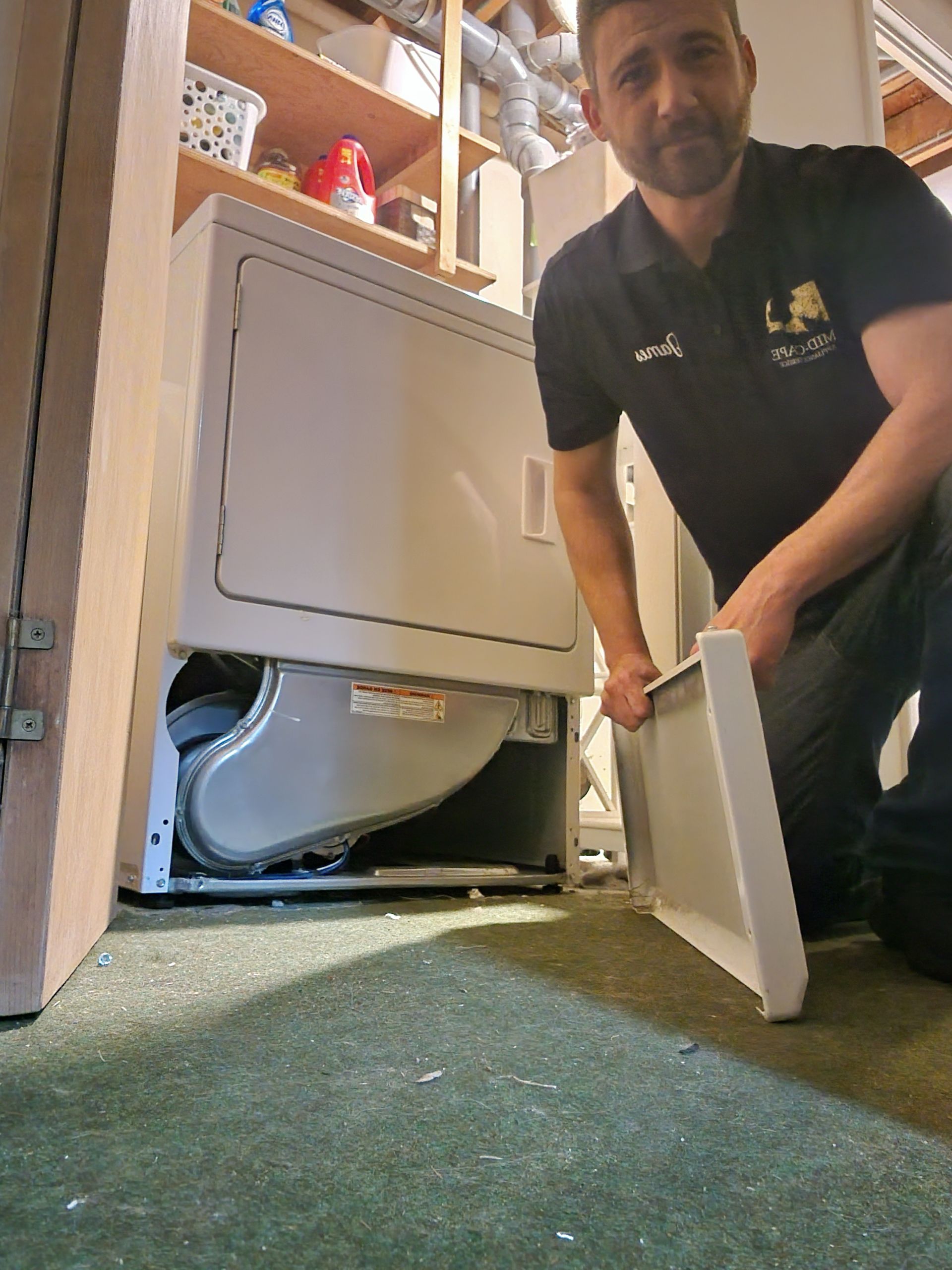 A technician holding a removed lower panel of a white dryer in a utility room.