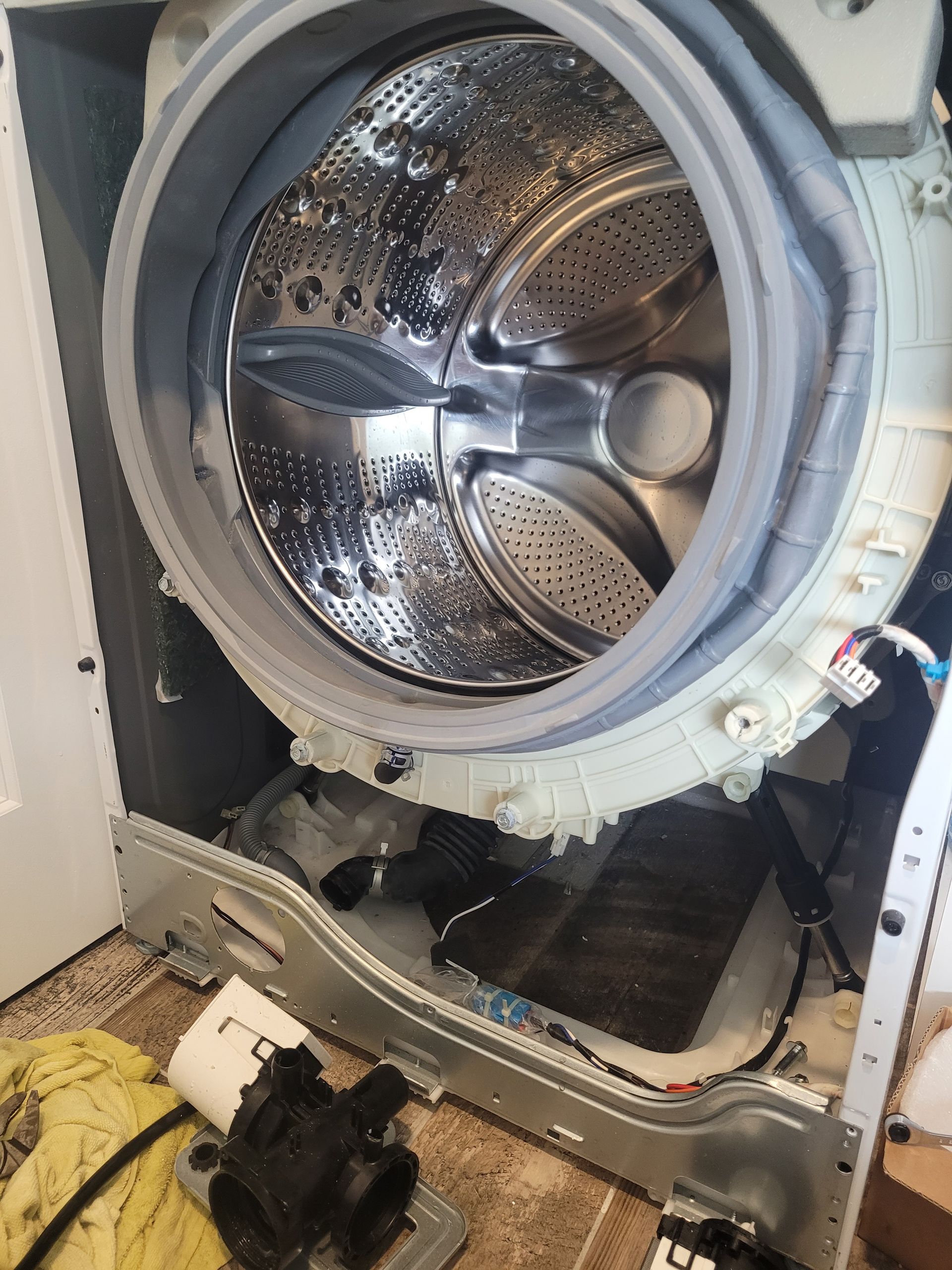 The front panel of a front-loading washing machine is removed, exposing the internal drum, white casing, and drain pump.