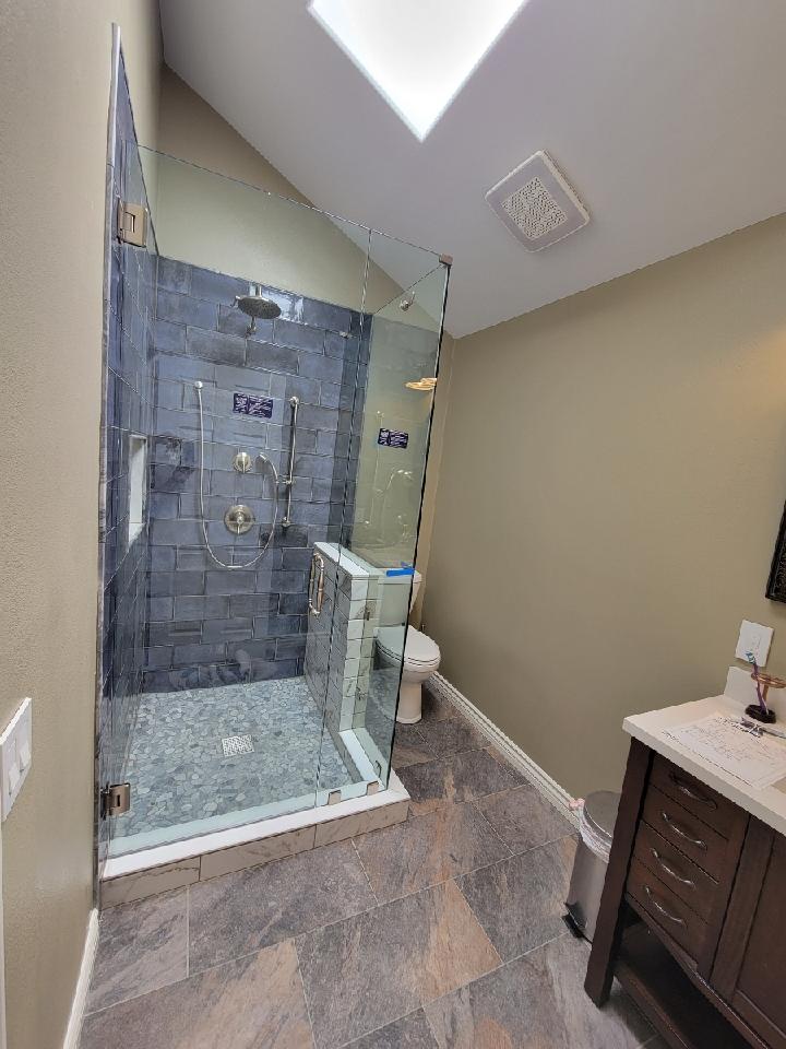 Bathroom with glass shower, blue-gray tile, white toilet, brown vanity, stone floor, and skylight.