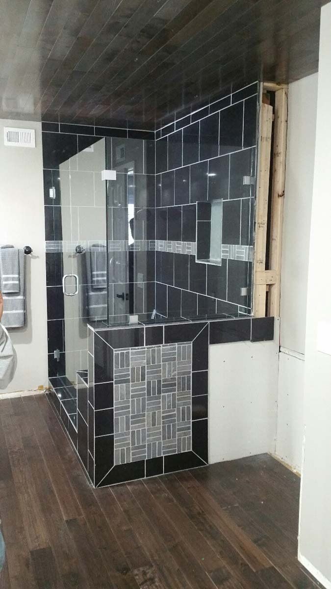 Black tiled shower with glass door, wood floor and unfinished wall section.