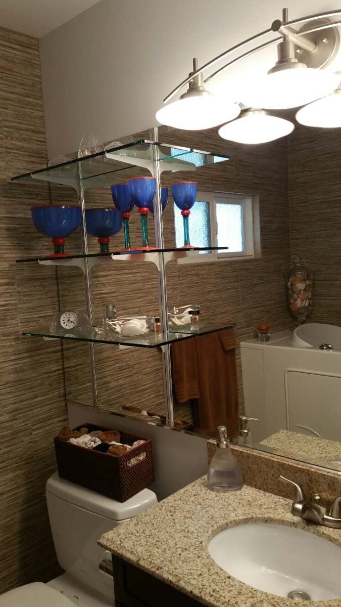 Bathroom with glass shelves holding blue glassware, above a toilet next to a sink with a granite countertop.