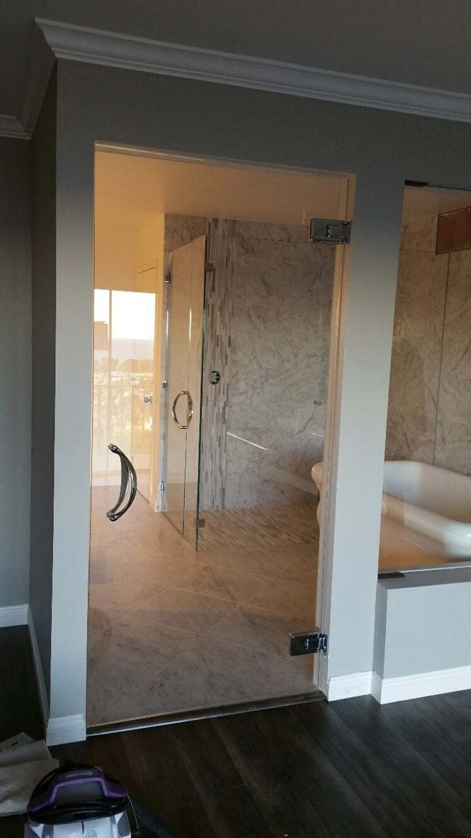 Glass shower door, hinges, and handle against gray walls, leading to a bathroom with a tub and toilet.