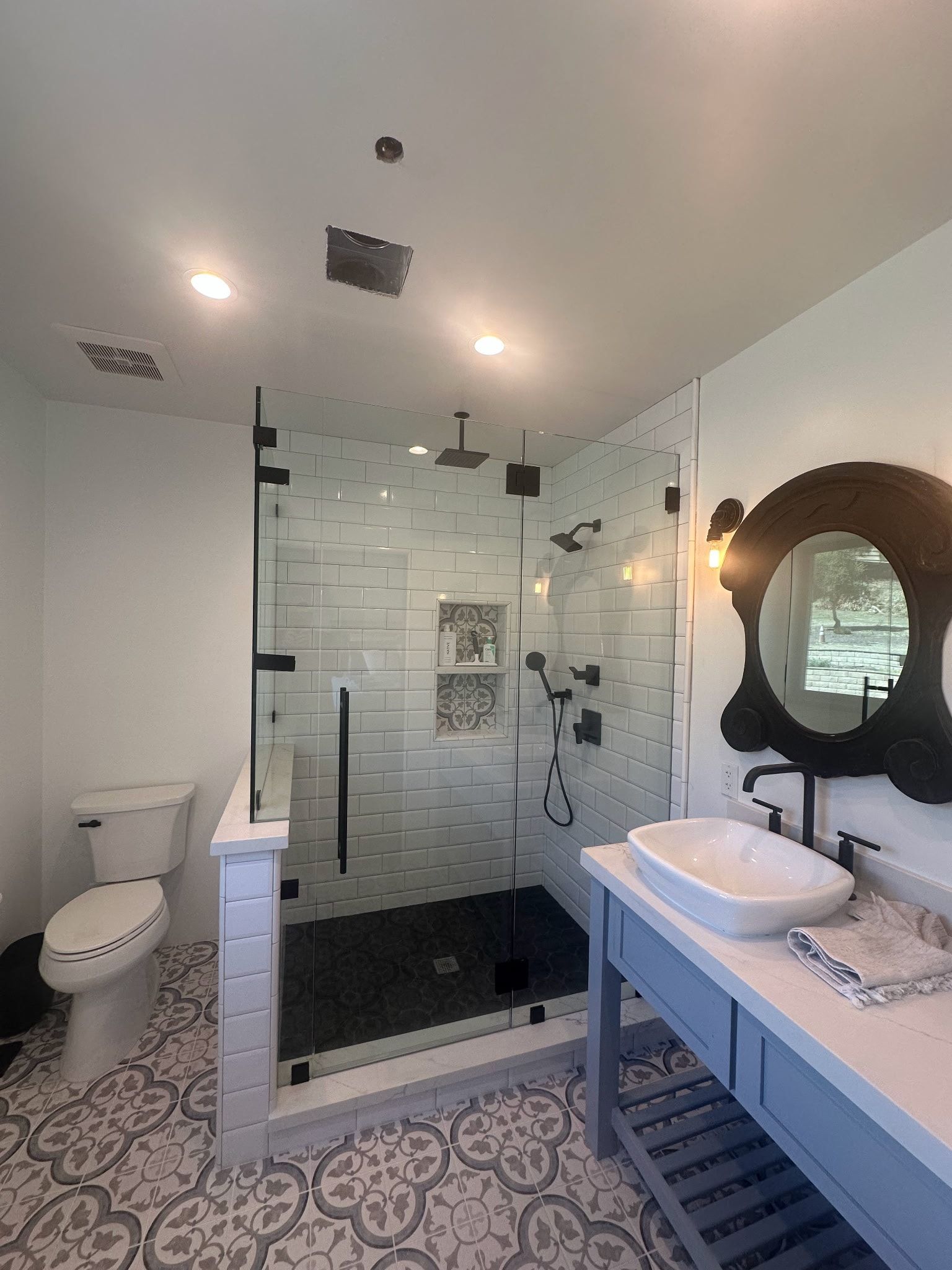 Bathroom with blue vanity, round mirror, glass shower with black fixtures, and patterned floor.