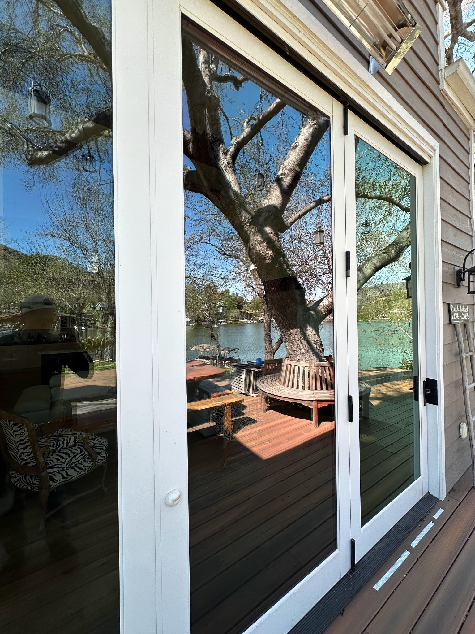 Glass patio doors reflecting a large tree, lake, and wooden deck. White frames, sunny day.