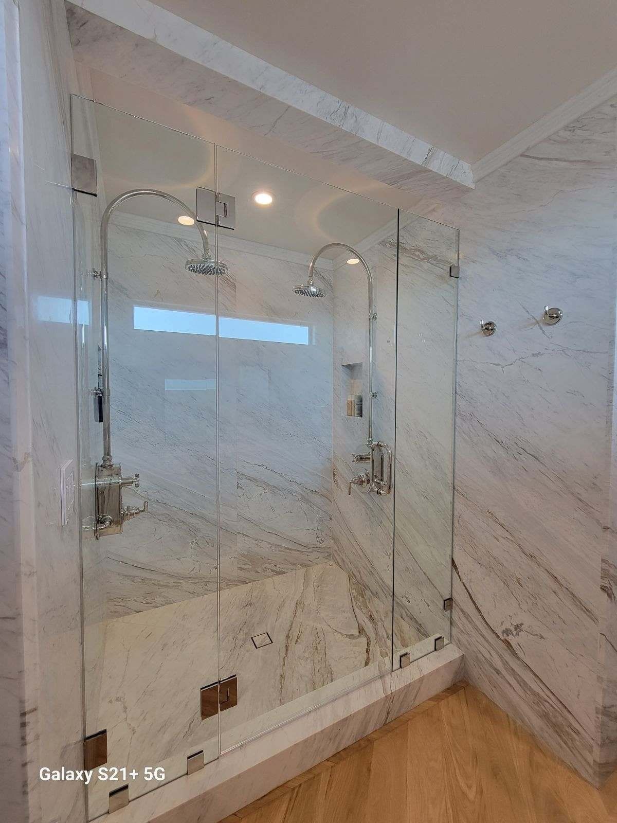 Glass shower enclosure with two showerheads, marble walls, and wood floor.