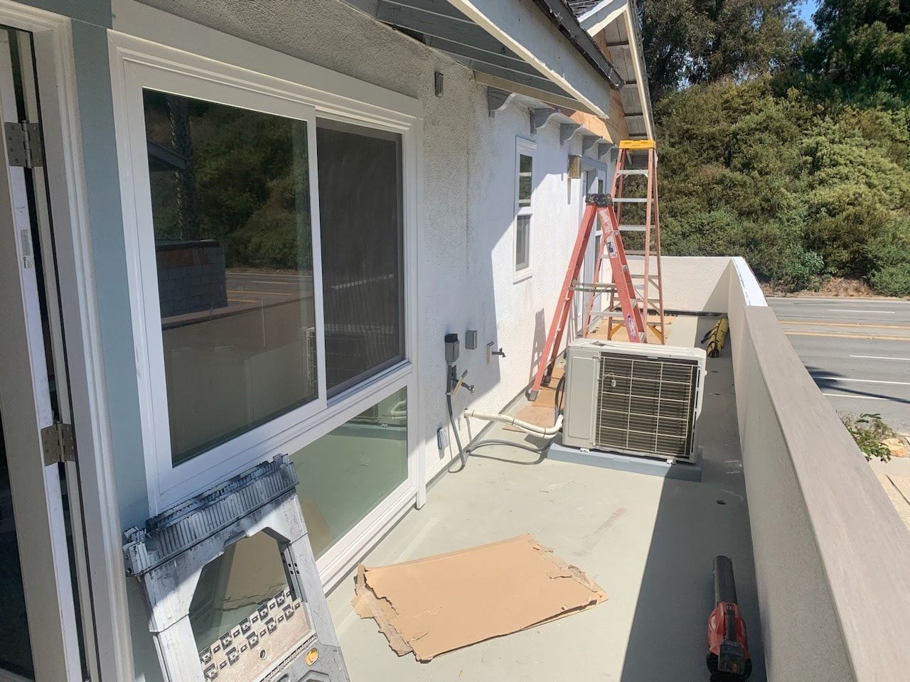 Balcony with a sliding glass door, a window, and an air conditioner unit, with a ladder in the background.