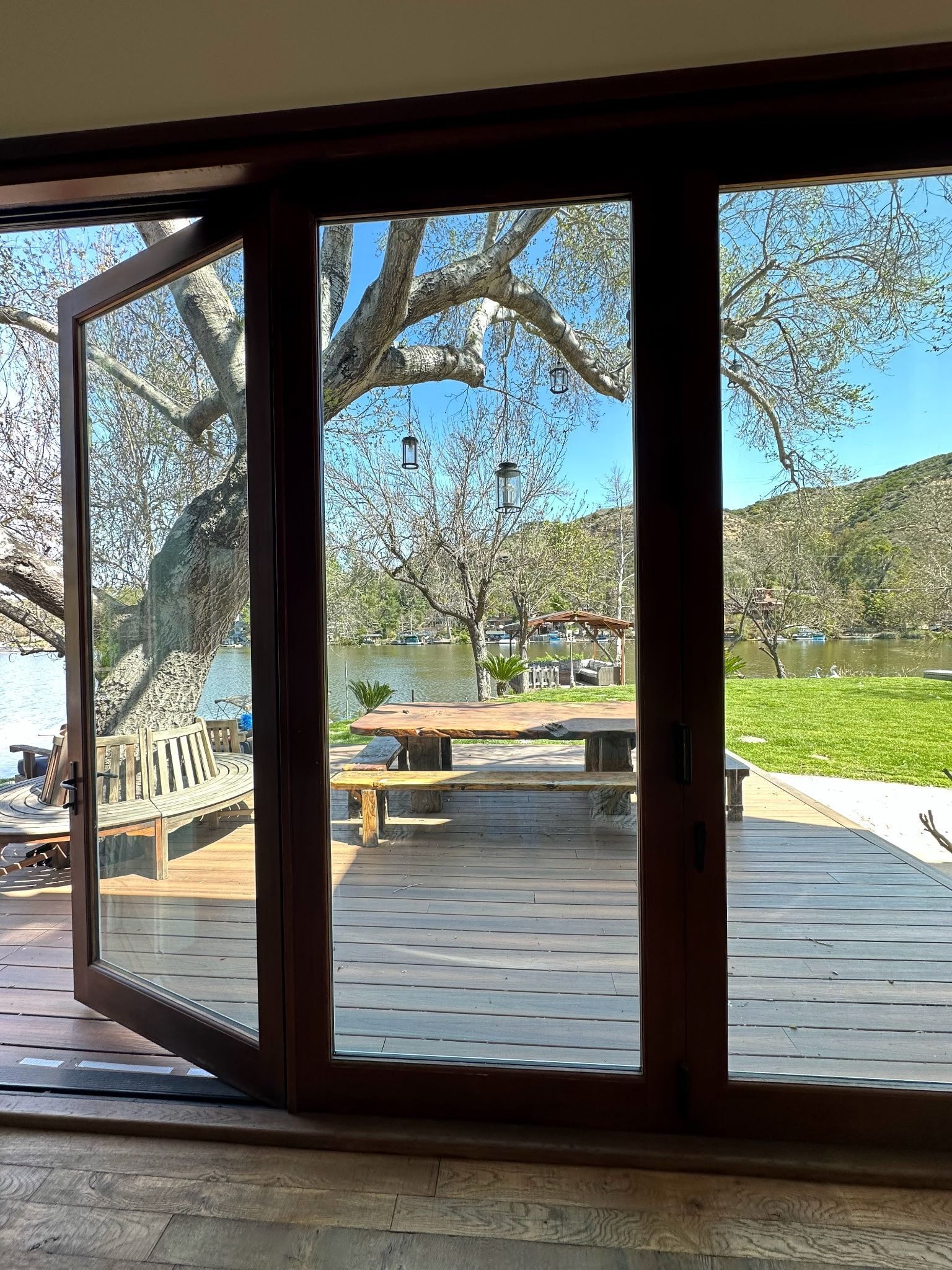 Wooden doors open onto a waterfront deck with a picnic table, tree, and green grass.