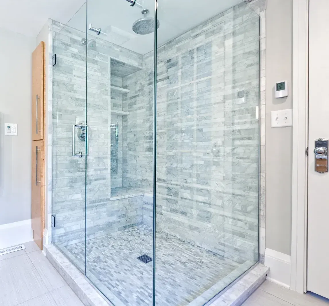 A corner shower with a glass door, light gray tile walls, and a pebble floor.