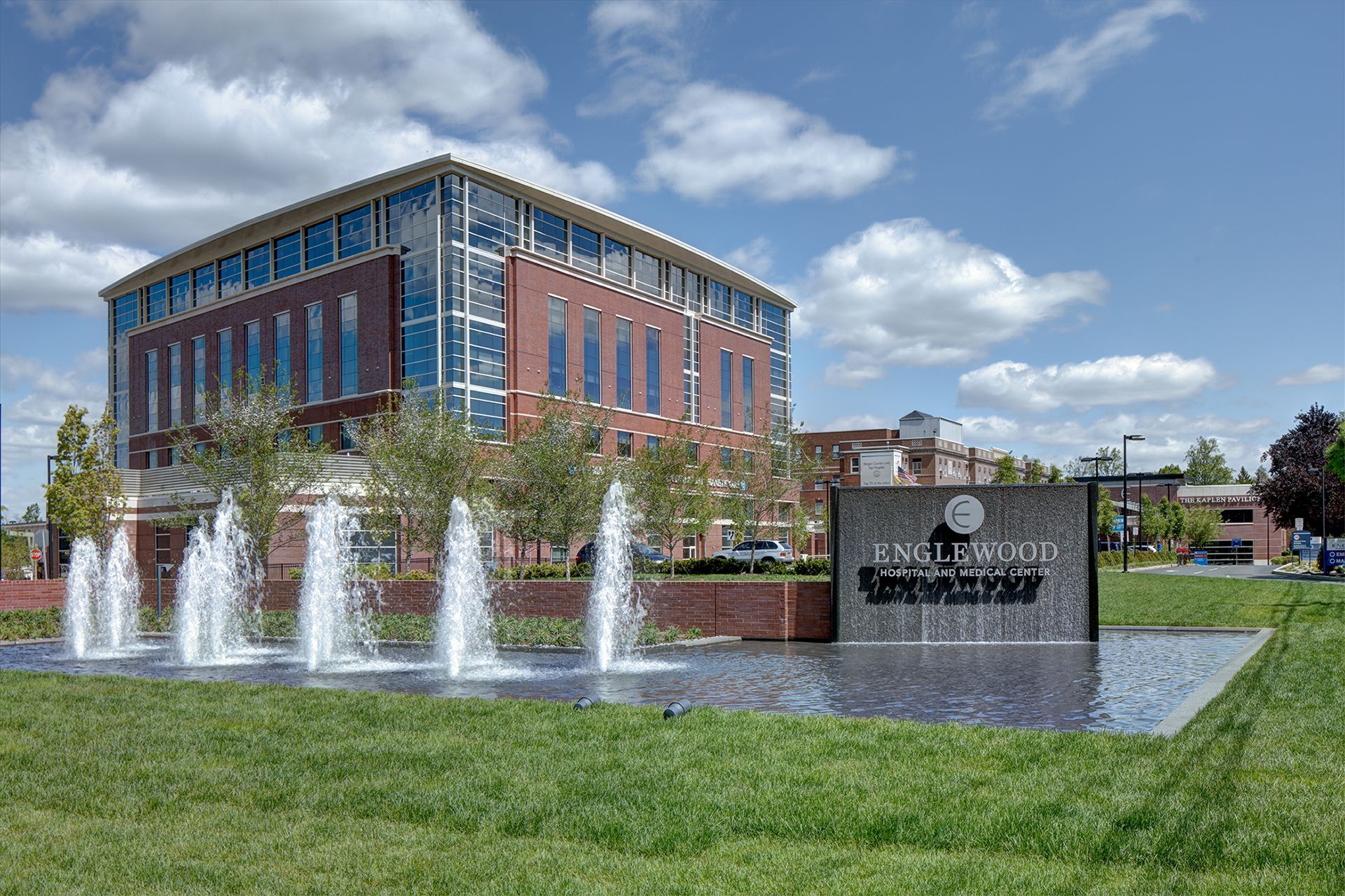 Fountain in front of a brick building with a glass facade, under a blue sky with clouds.