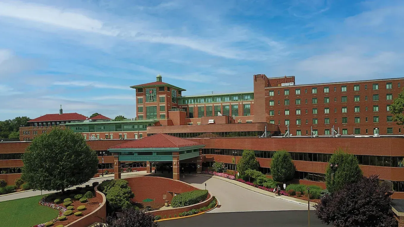 Large brick building with a green roof and a circular drive. Blue sky in the background.