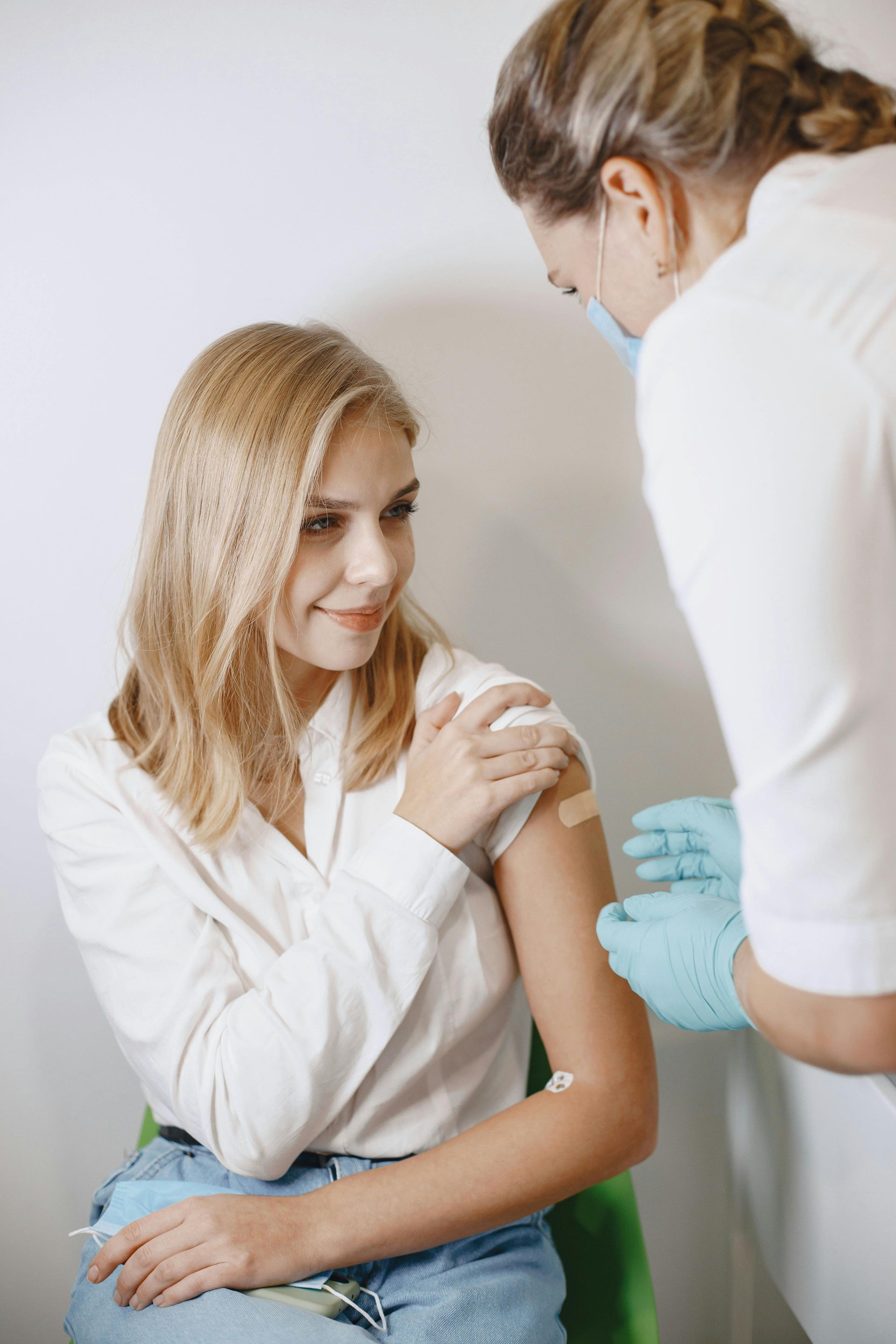 Woman getting a vaccine shot from a person in a medical setting; she smiles, covering her arm with a bandage.