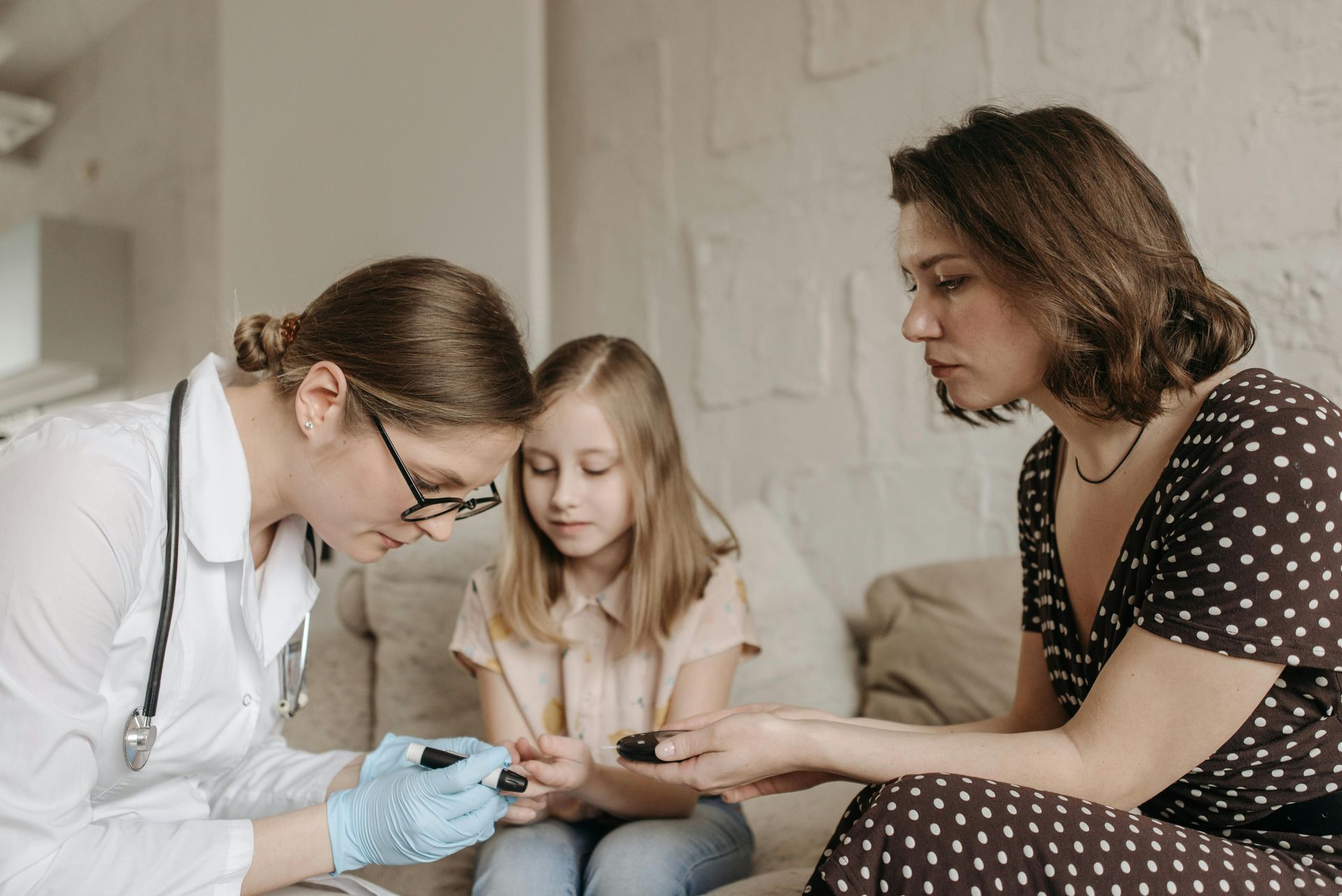 Doctor checking a child's finger with a blood glucose monitor, mother watches, indoor setting.