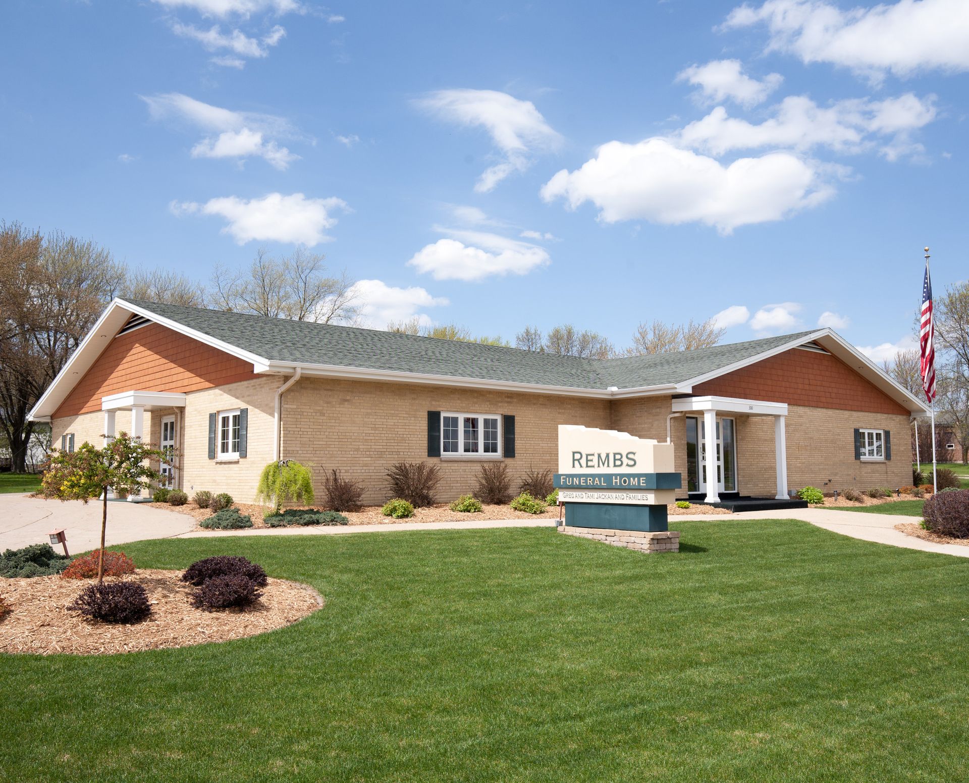 Exterior of a light brown brick building with a green lawn, sign, and American flag. 