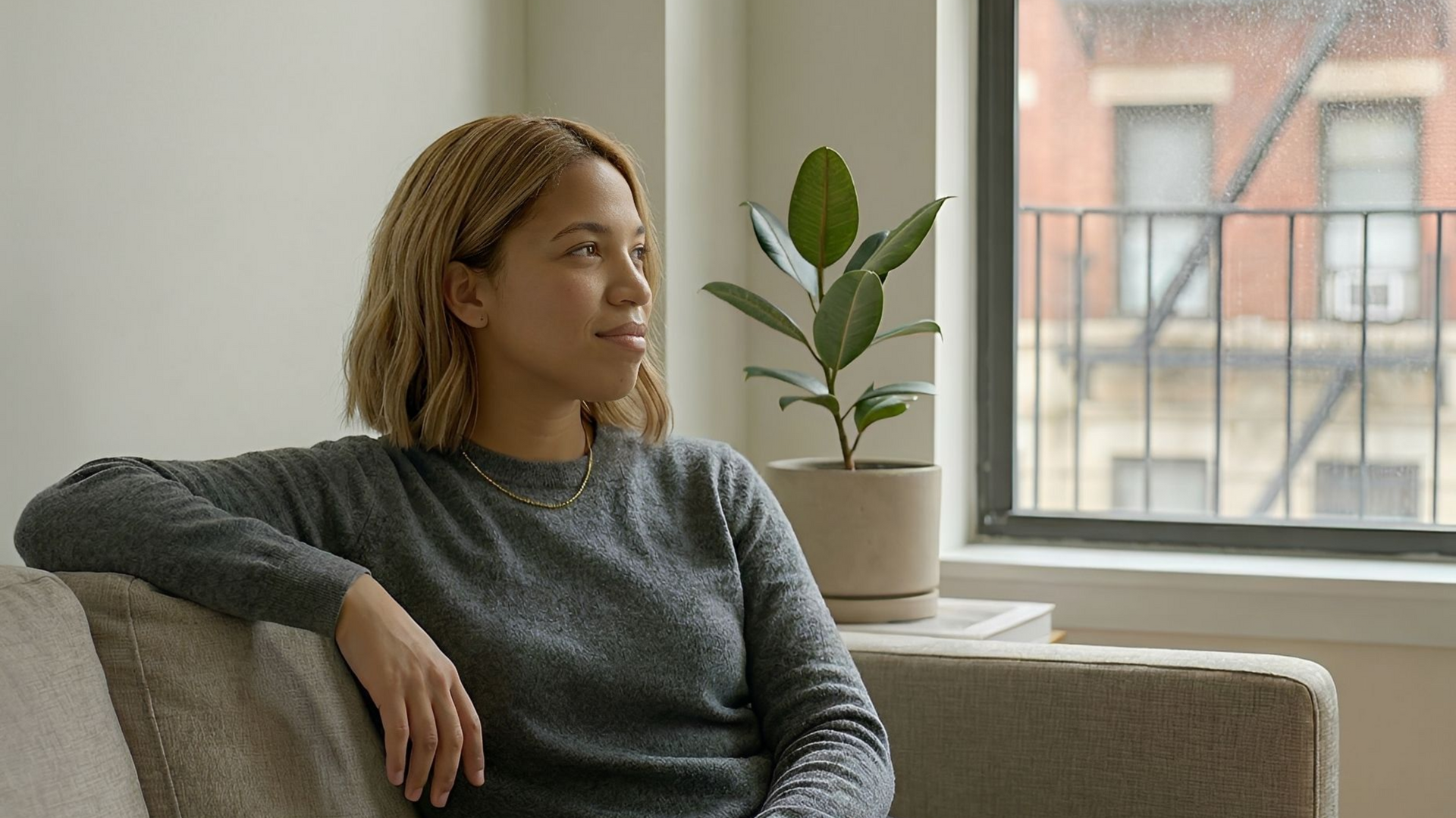 Woman in gray sweater looks out window while sitting on a couch next to a plant.