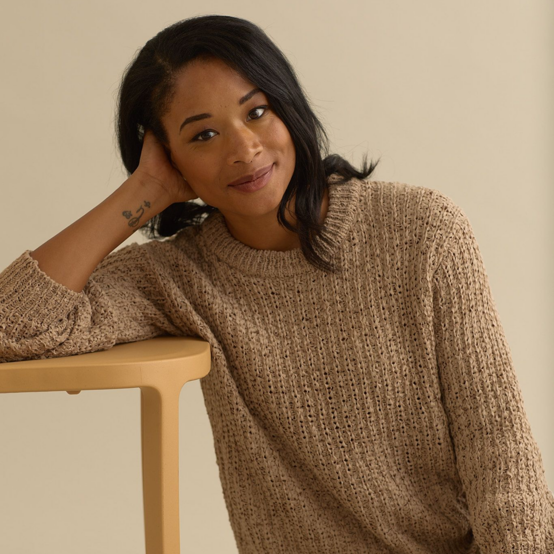 Woman leaning on a small table, wearing a beige sweater, smiling. Light-colored backdrop.