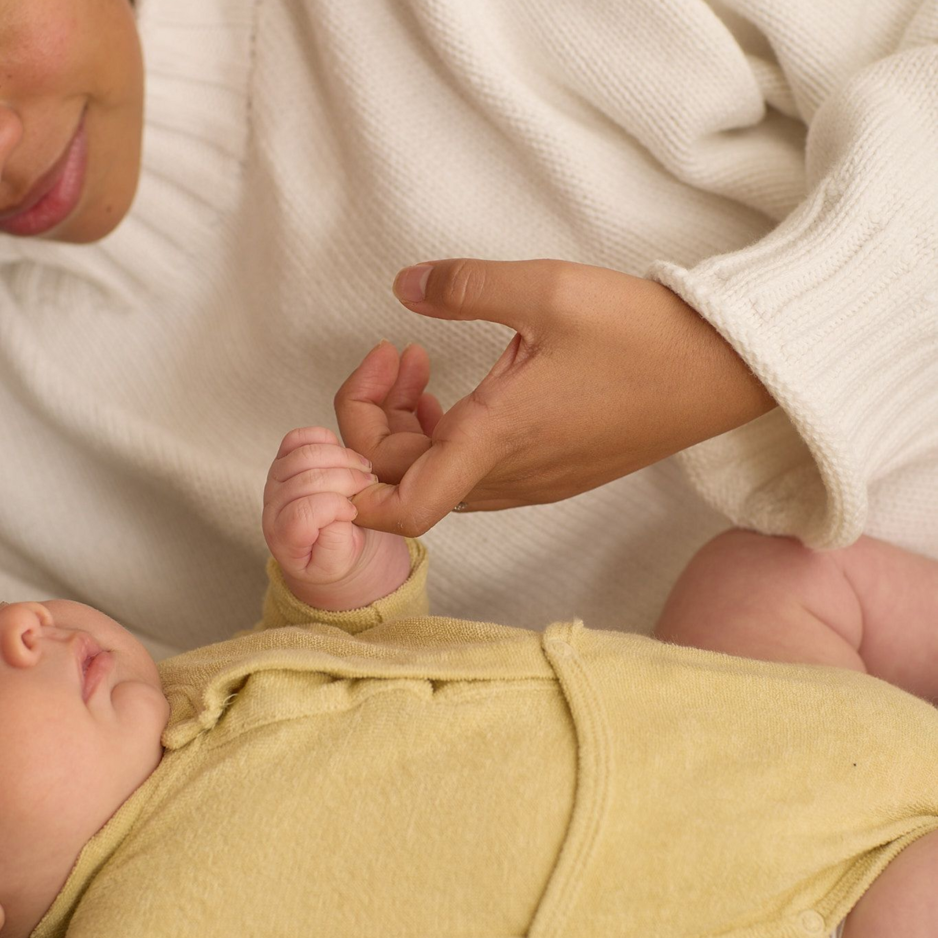 Baby's hand clasped by an adult's finger; baby wearing a yellow onesie, adult in a white sweater.