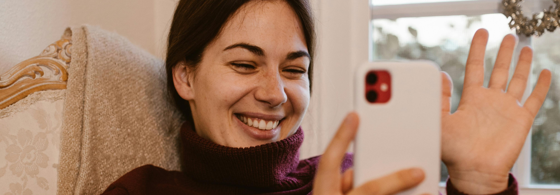 Woman smiles while video chatting on her phone, waving at the screen. She is wearing a dark turtleneck and seated indoors.