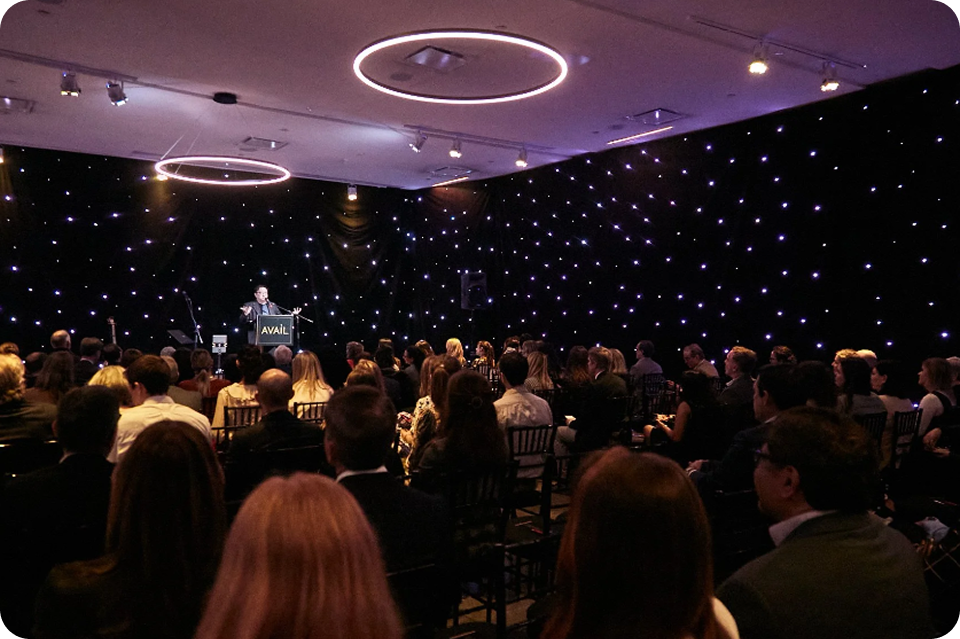 Audience in a dark room with a speaker at a podium under stage lights, with a starry backdrop.